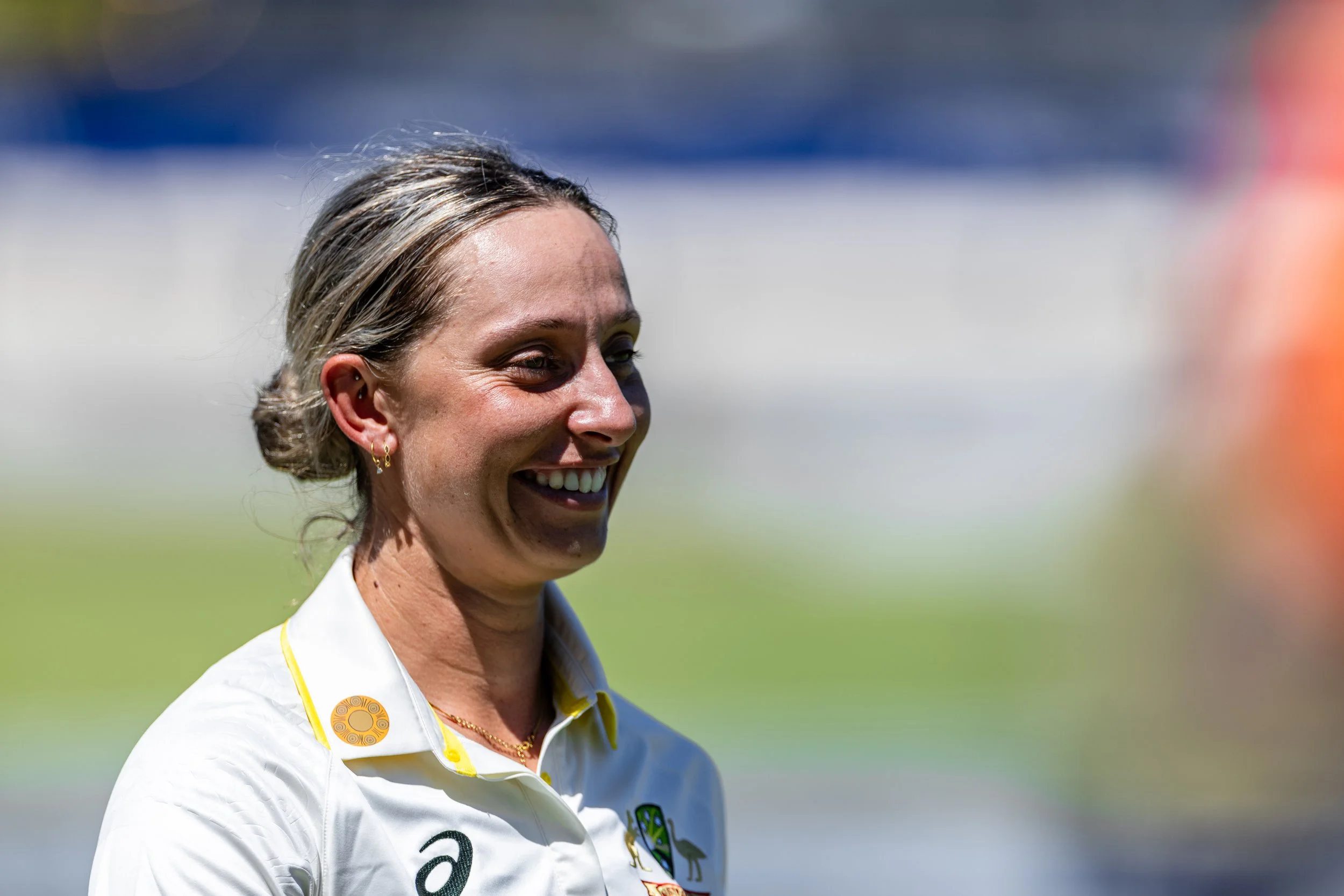 A woman smiling outdoors, wearing a white shirt with yellow accents, gold earrings, and a necklace, with a blurred background of green grass and sky.