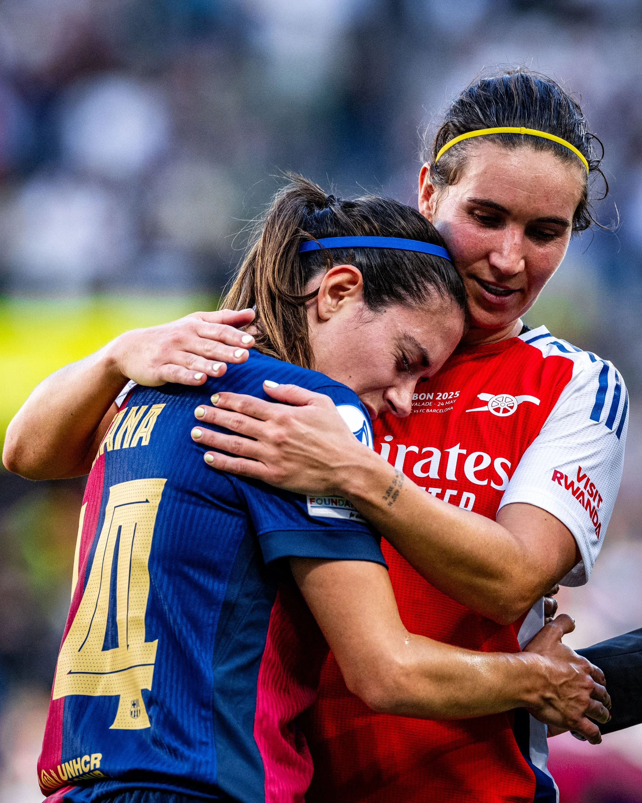 Two female soccer players hug in celebration on the field. One is wearing a blue jersey with yellow accents and the number 14, the other is wearing a red and white jersey with text referencing Emirates and a date, with a yellow headband. The backgrou