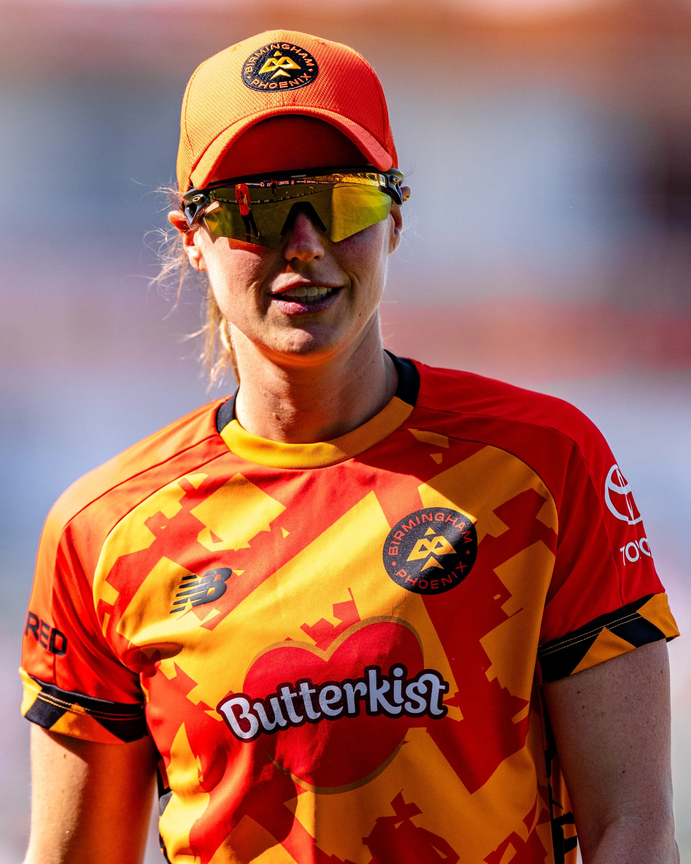 Close-up of a female athlete wearing a red and yellow sports jersey with the word '#Butterkist' on it, an orange cap with a logo, and reflective sunglasses, outdoors during a sporting event.