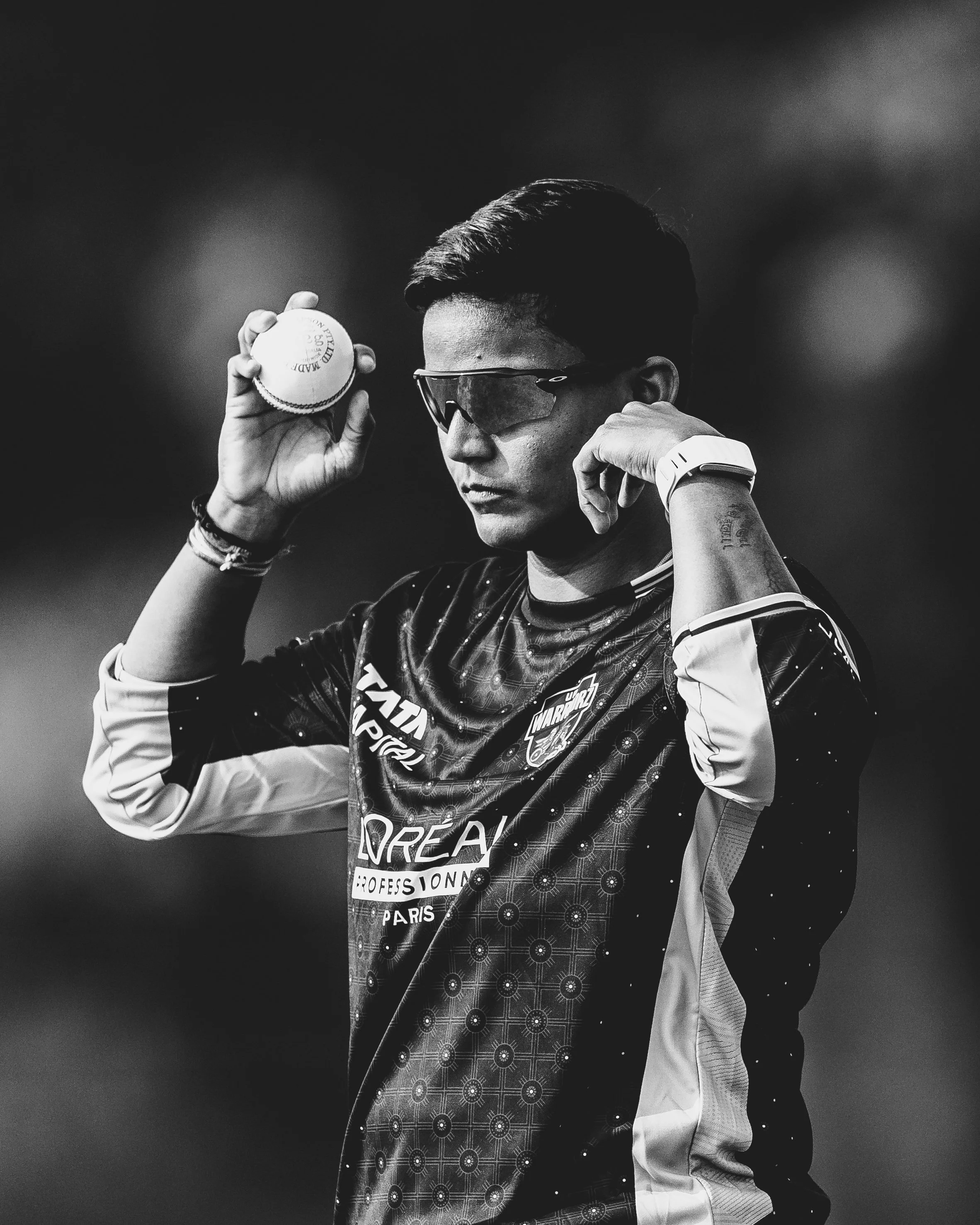 A young male athlete holding a tennis ball to his ear, wearing sunglasses and sports attire, with a serious expression.