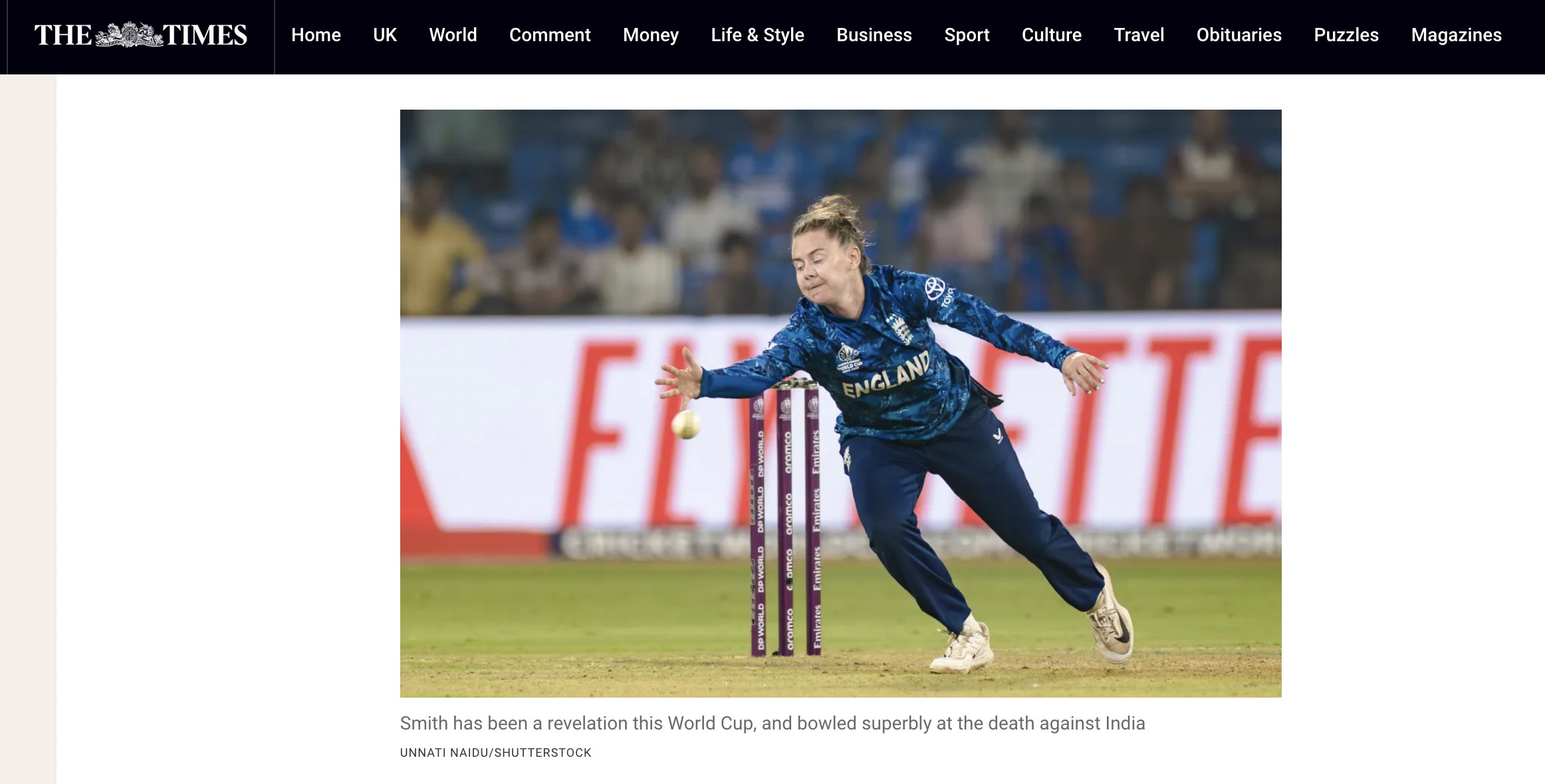 A female athlete in a blue England sports jacket is bowling with her right hand during a cricket match. The background is a stadium with blurred spectators, and the cricket stumps are visible.