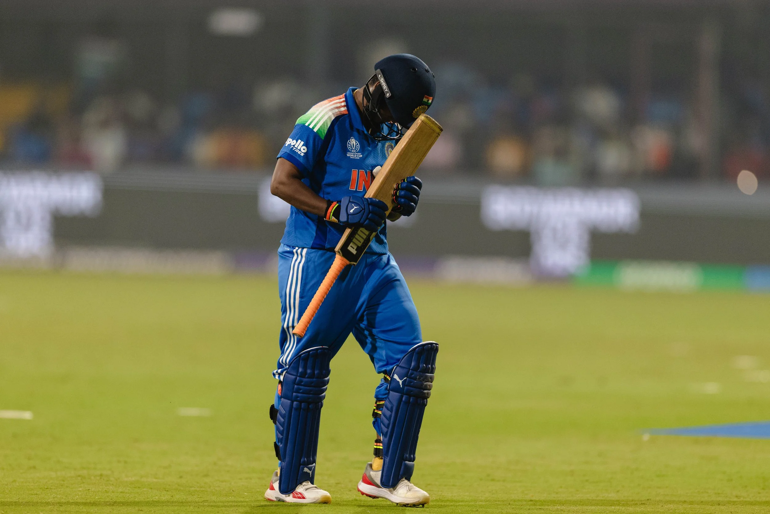 An Indian cricket player dressed in blue pads, jersey, and helmet, walking on the cricket field while holding a cricket bat.