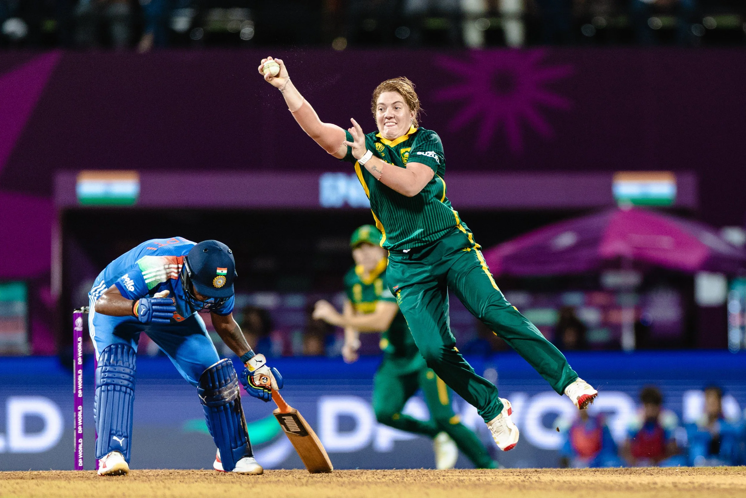 A female cricket player in a green and yellow uniform leaps into the air to hit a cricket ball during a match, with a male umpire in blue crouched near the stumps.