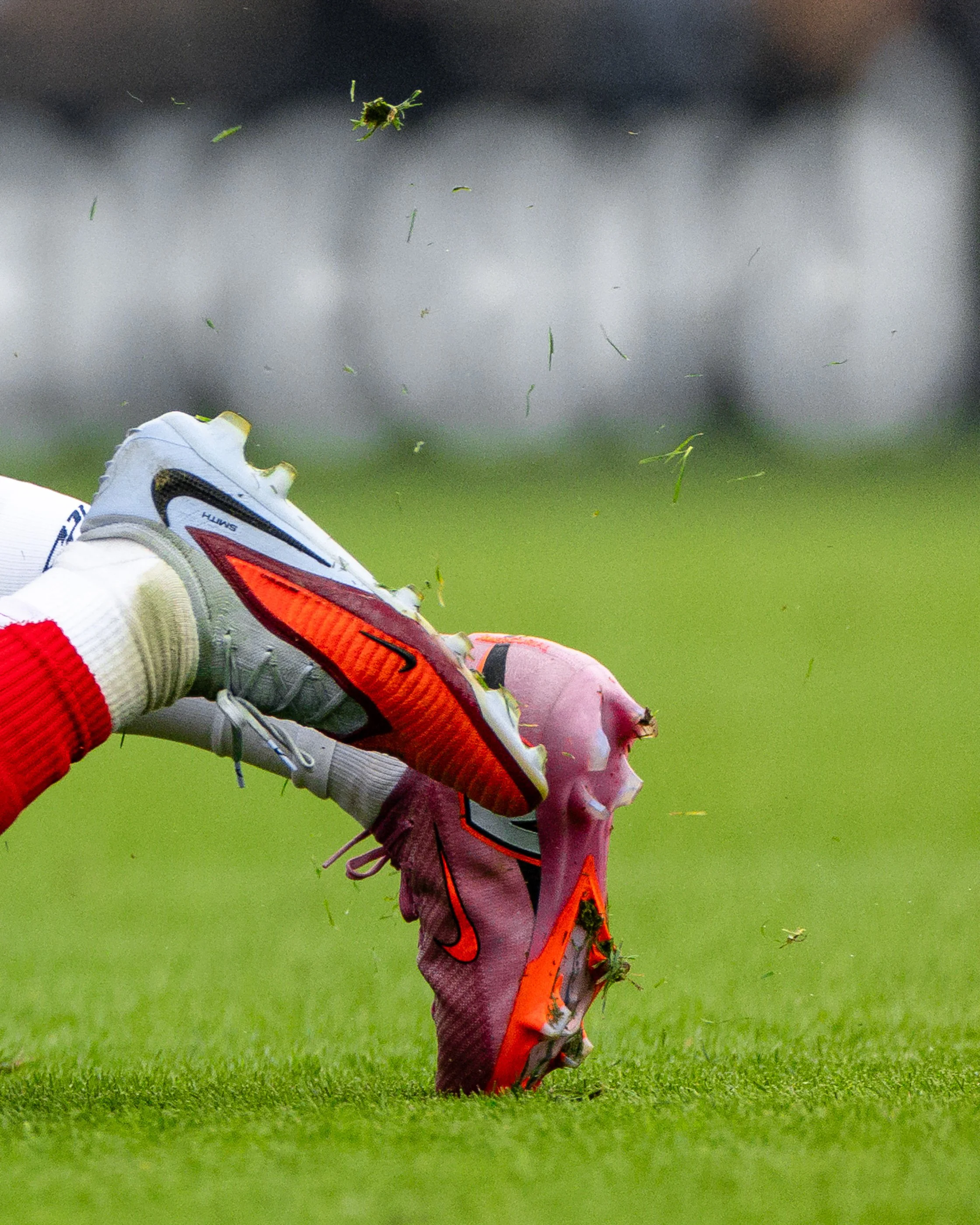 Close-up view of a person's foot wearing a white athletic shoe with orange accents kicking a pink tennis ball on a grassy field, with small green debris flying around.