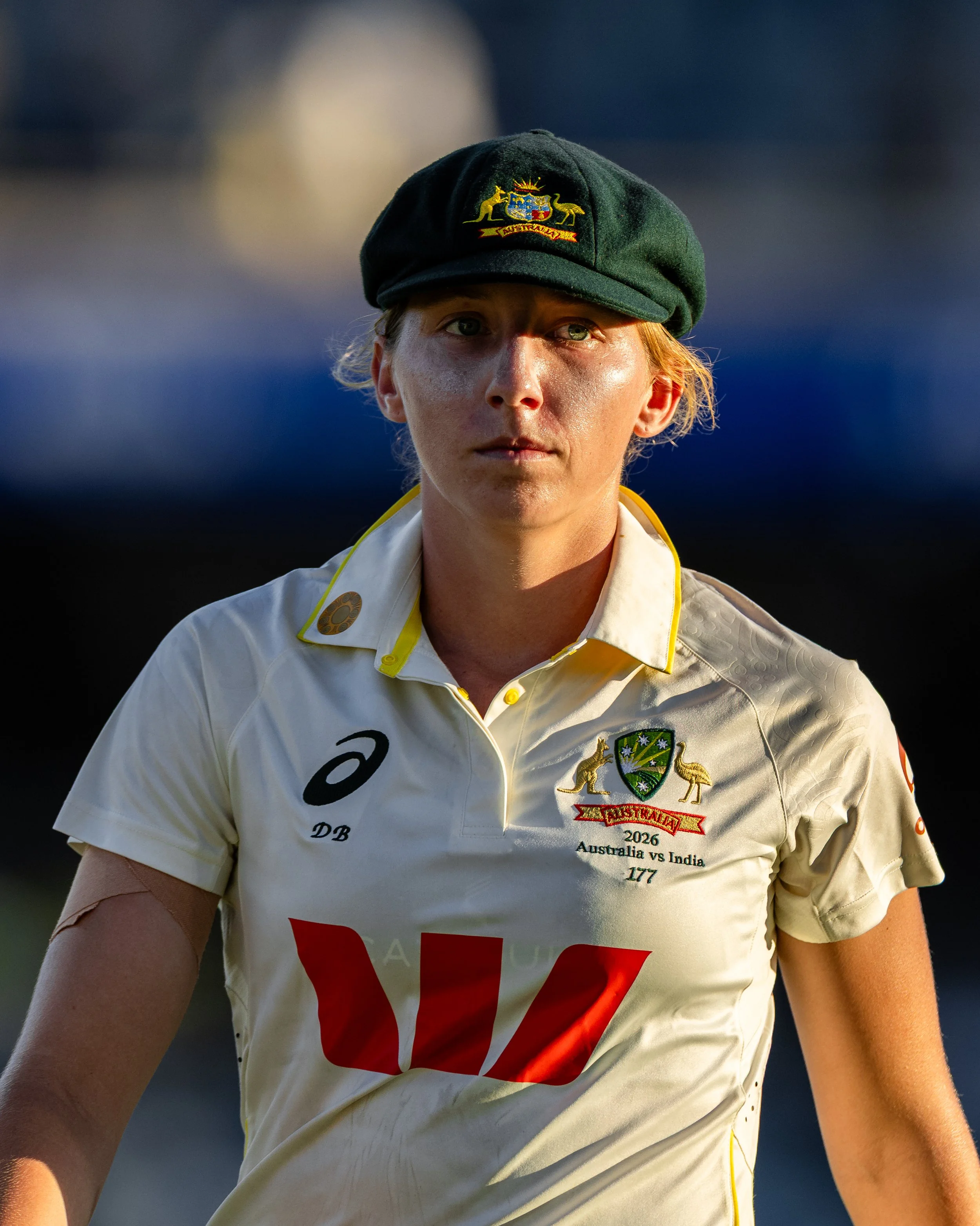 Close-up of a female cricket player from Australia wearing a white uniform and a dark green cap with the Australian coat of arms. The uniform features sponsorship logos and text including 'Australia vs India 2026.' The background is blurred, emphasiz