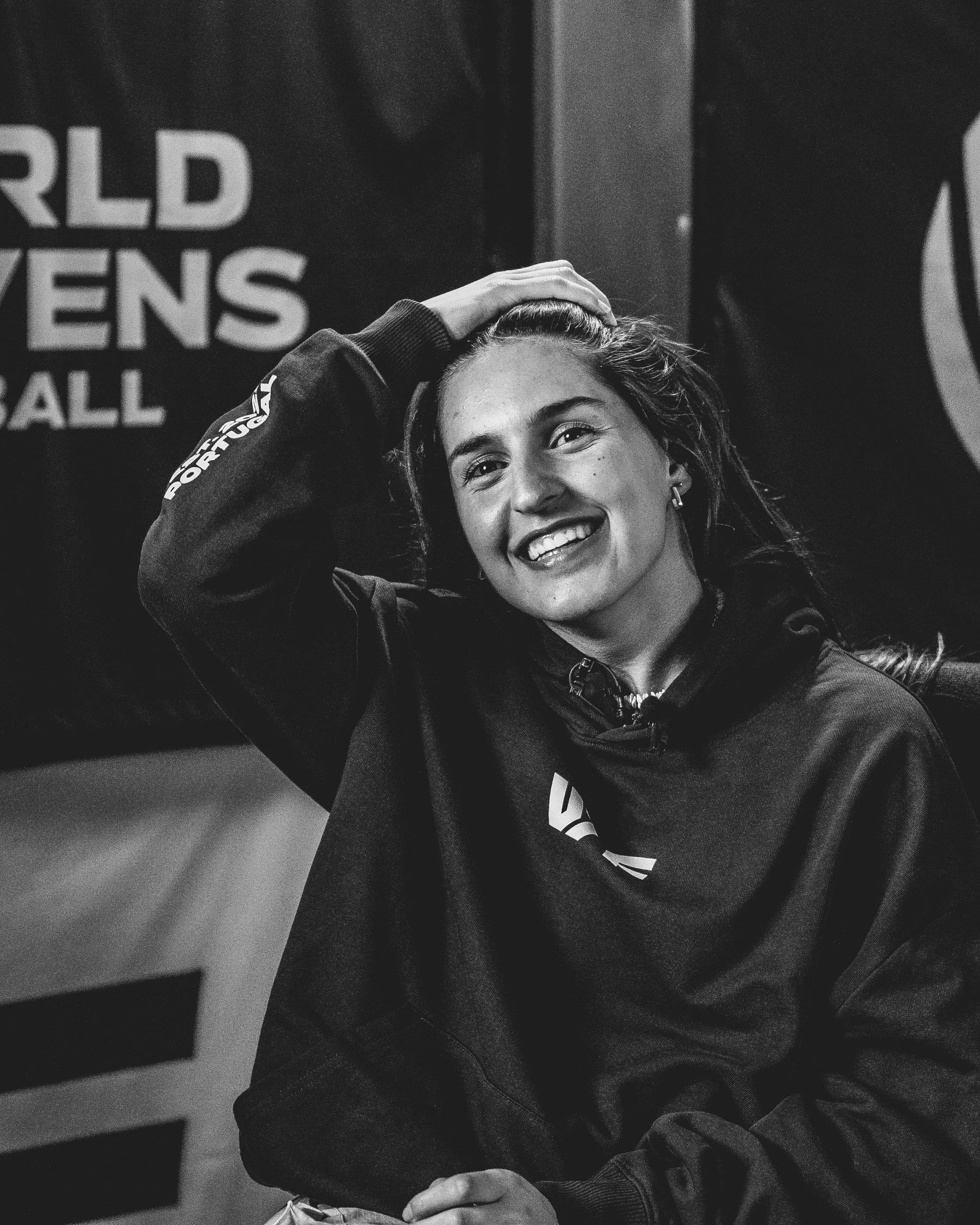 A young woman with a bright smile, holding her head with one hand, wearing a dark jacket with a Superman logo, in front of a backdrop with partially visible text and logos.