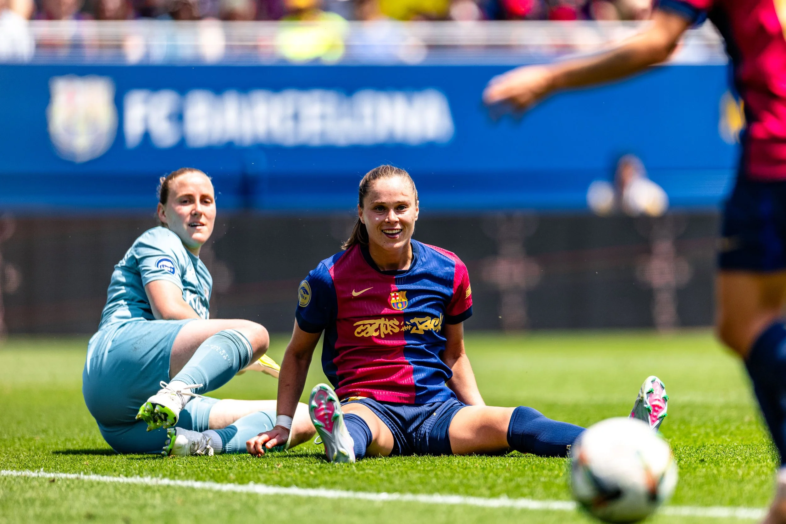 Two female soccer players, one in blue and the other in red and blue, sitting on the grass after a fall during a match, with a blurred background of spectators and a blue advertisement banner.