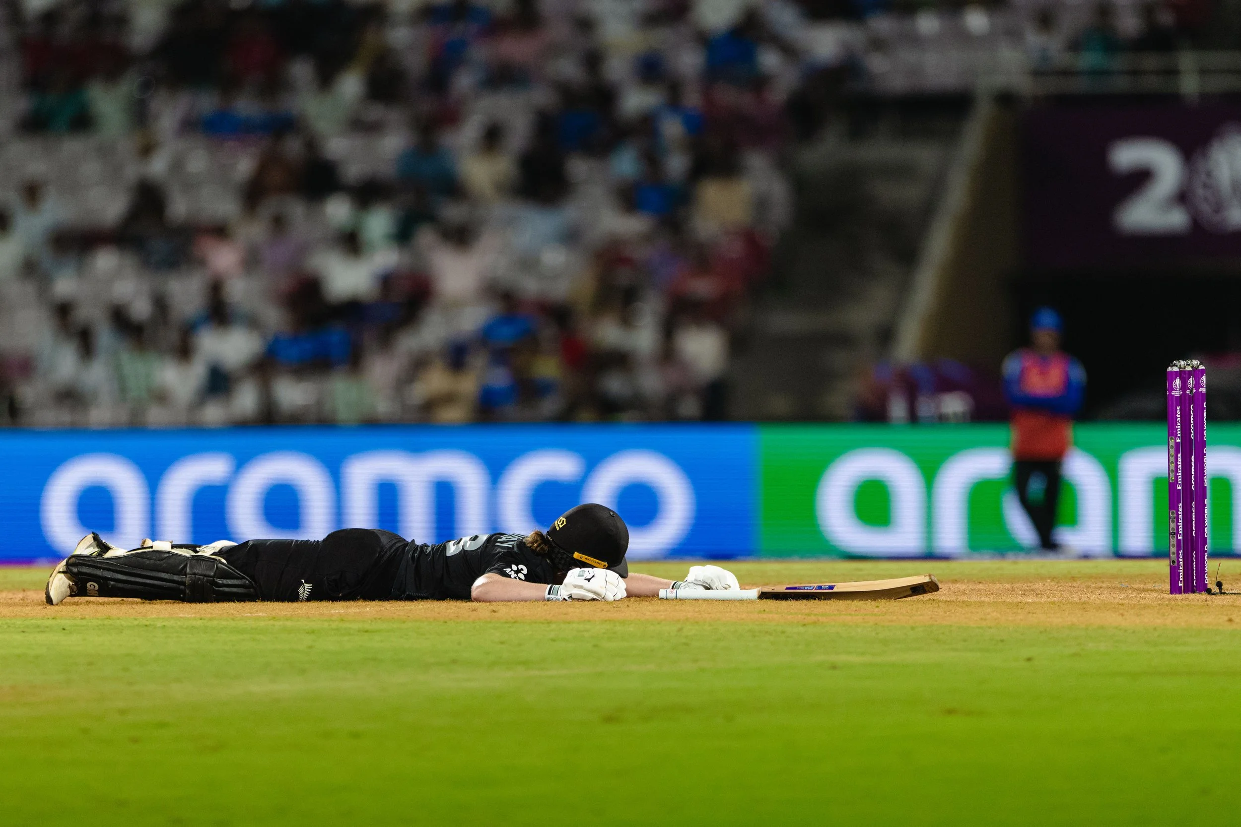 Cricket player lying on the field after being dismissed, with wickets and a blurred crowd in the background.
