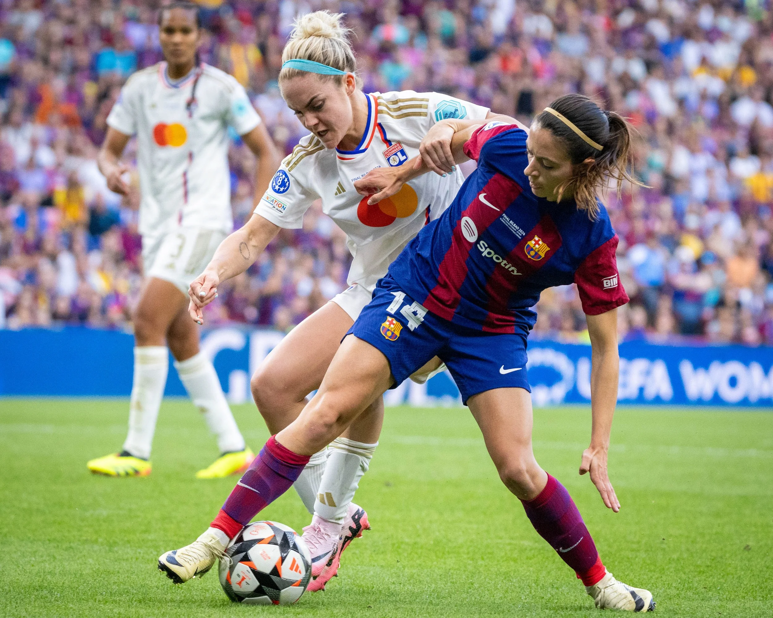 Two women playing soccer compete for the ball during a match, with a crowd in the background.