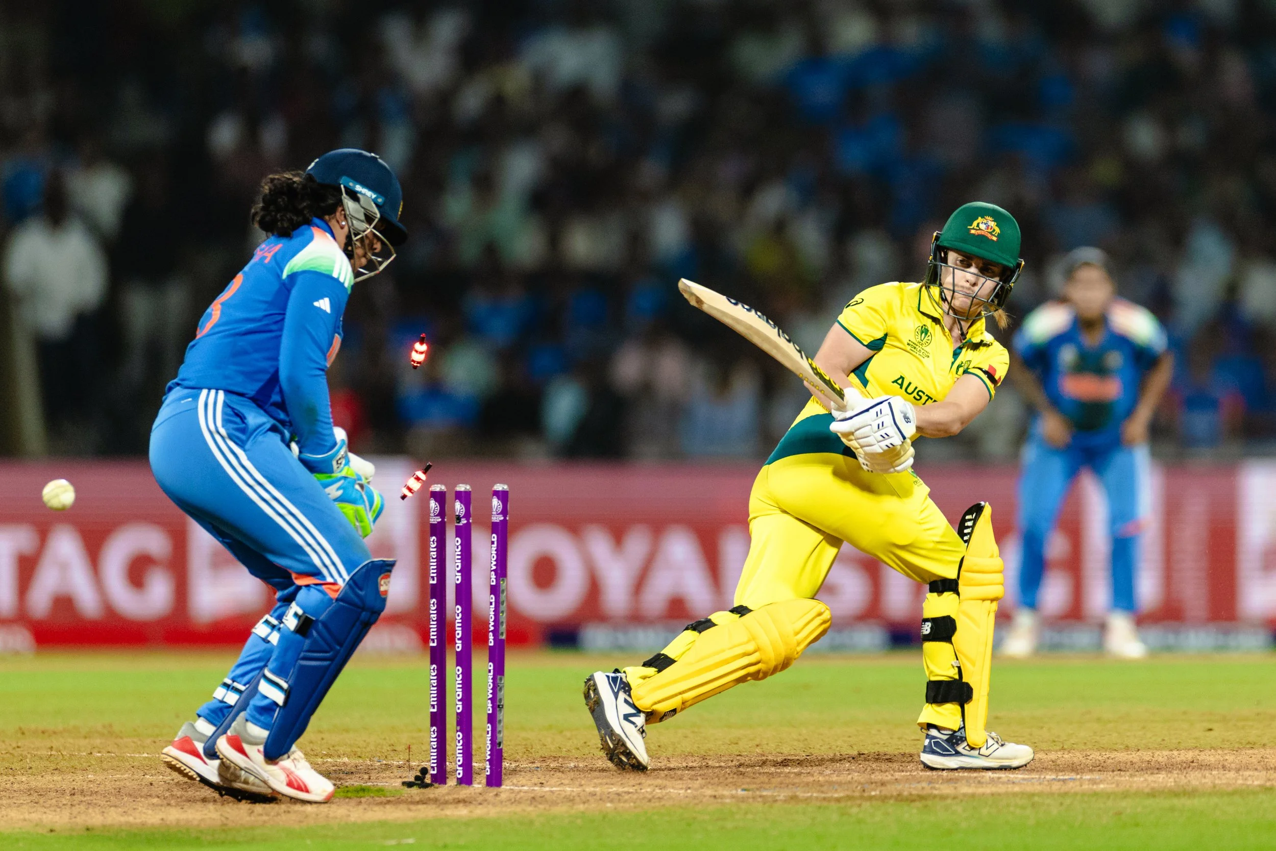 Women's cricket match with Australian batter trying to hit the ball while Indian wicketkeeper crouches to catch it, crowd in the background.