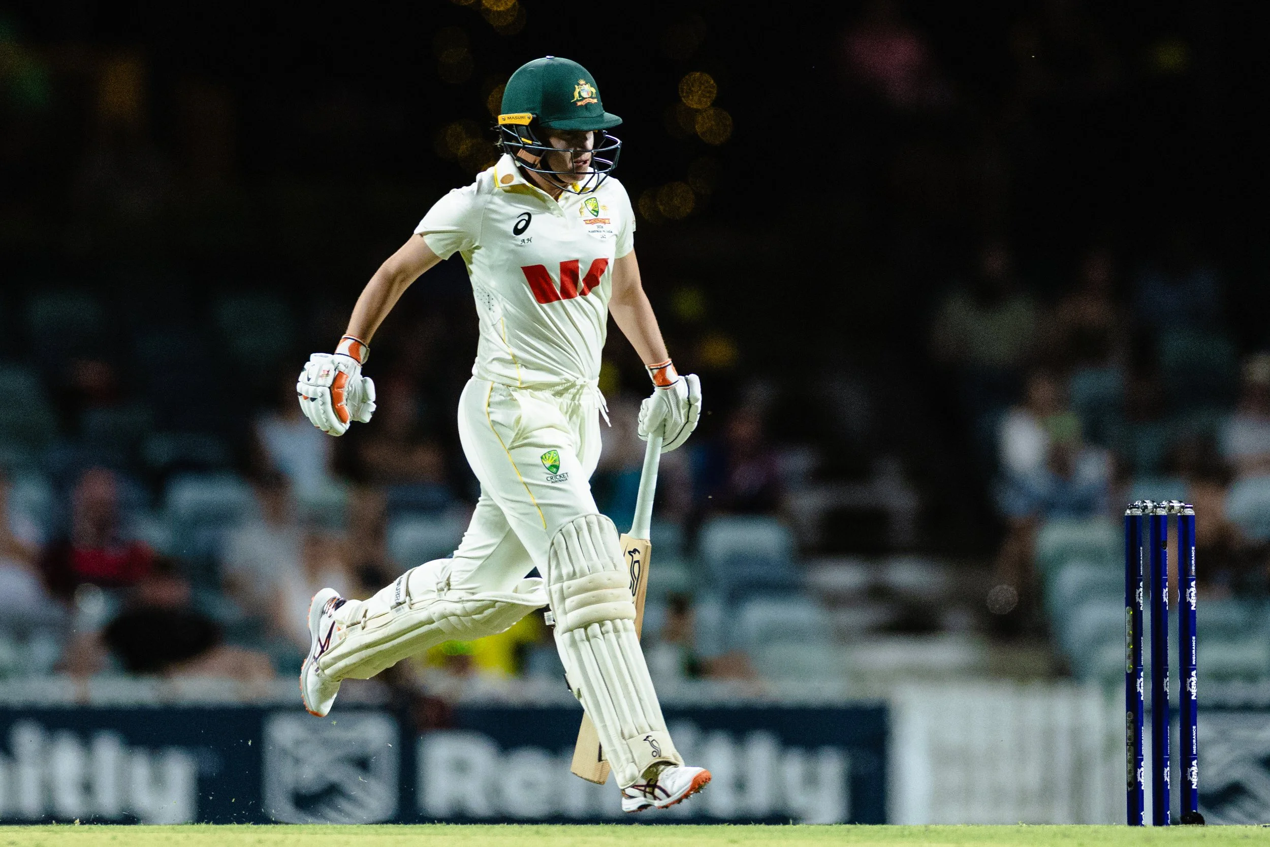 Cricket player celebrating on the field at night, wearing a green helmet and white uniform.