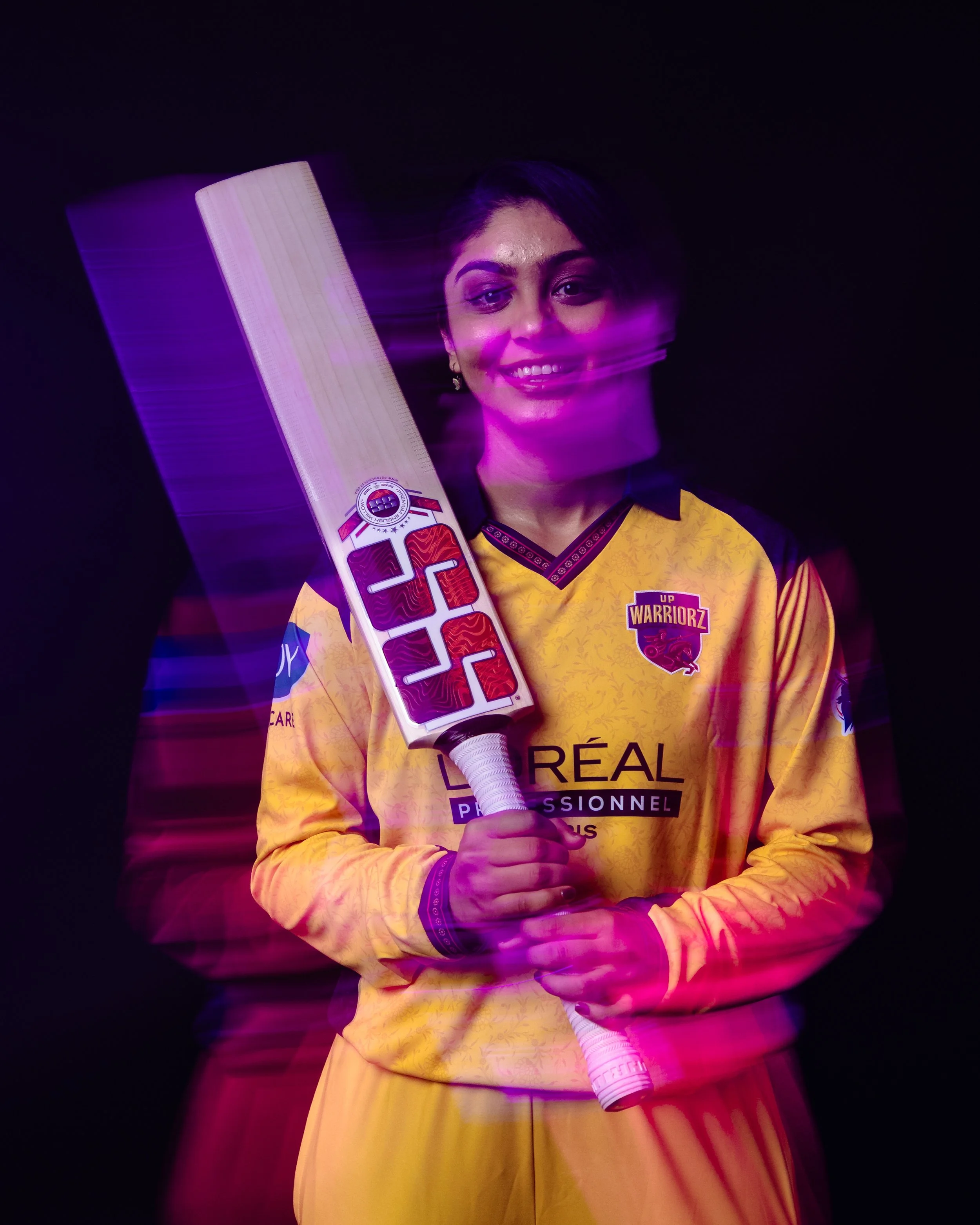 A woman with short dark hair and earrings is smiling while holding a cricket bat with her right hand. She is wearing a yellow sports jersey with black and red accents, and black lettering that reads 'UP WARRIORZ.' The background is black, and purple 