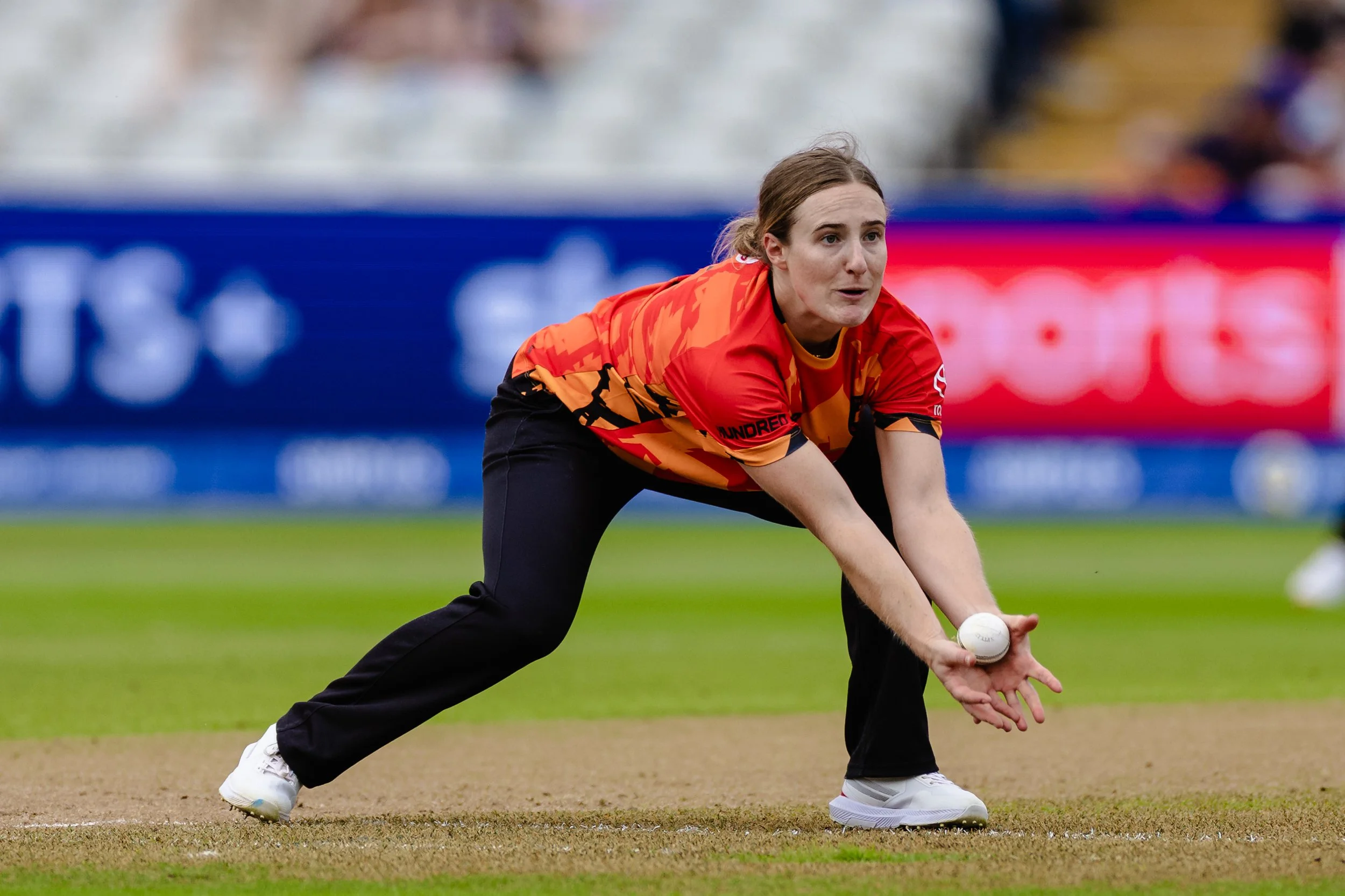 A female cricket player in a red and orange uniform bends down to pick up a cricket ball on the field.