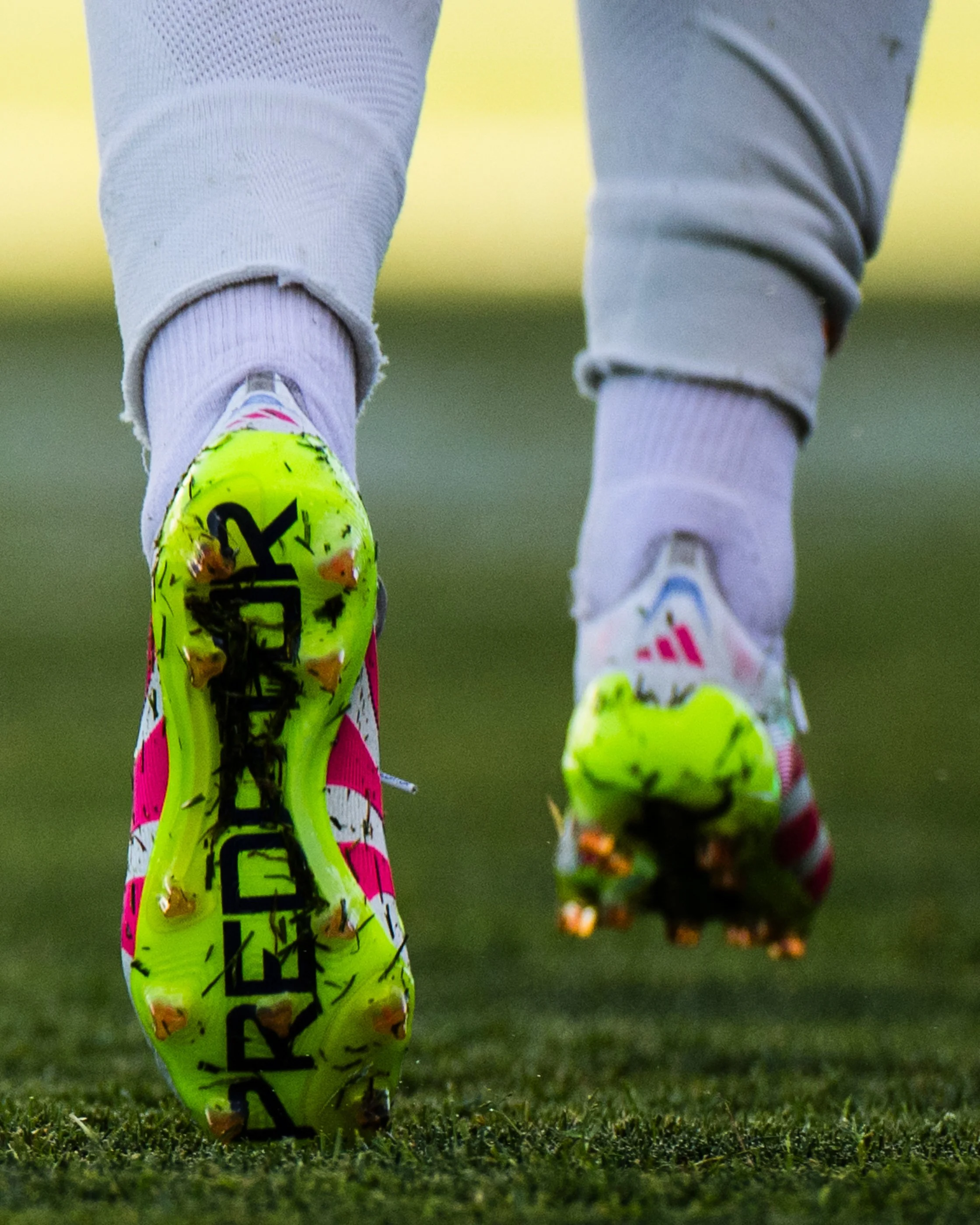 Close-up of a soccer player's foot wearing a bright yellow cleat with black and pink accents, kicking a soccer ball on a grassy field.