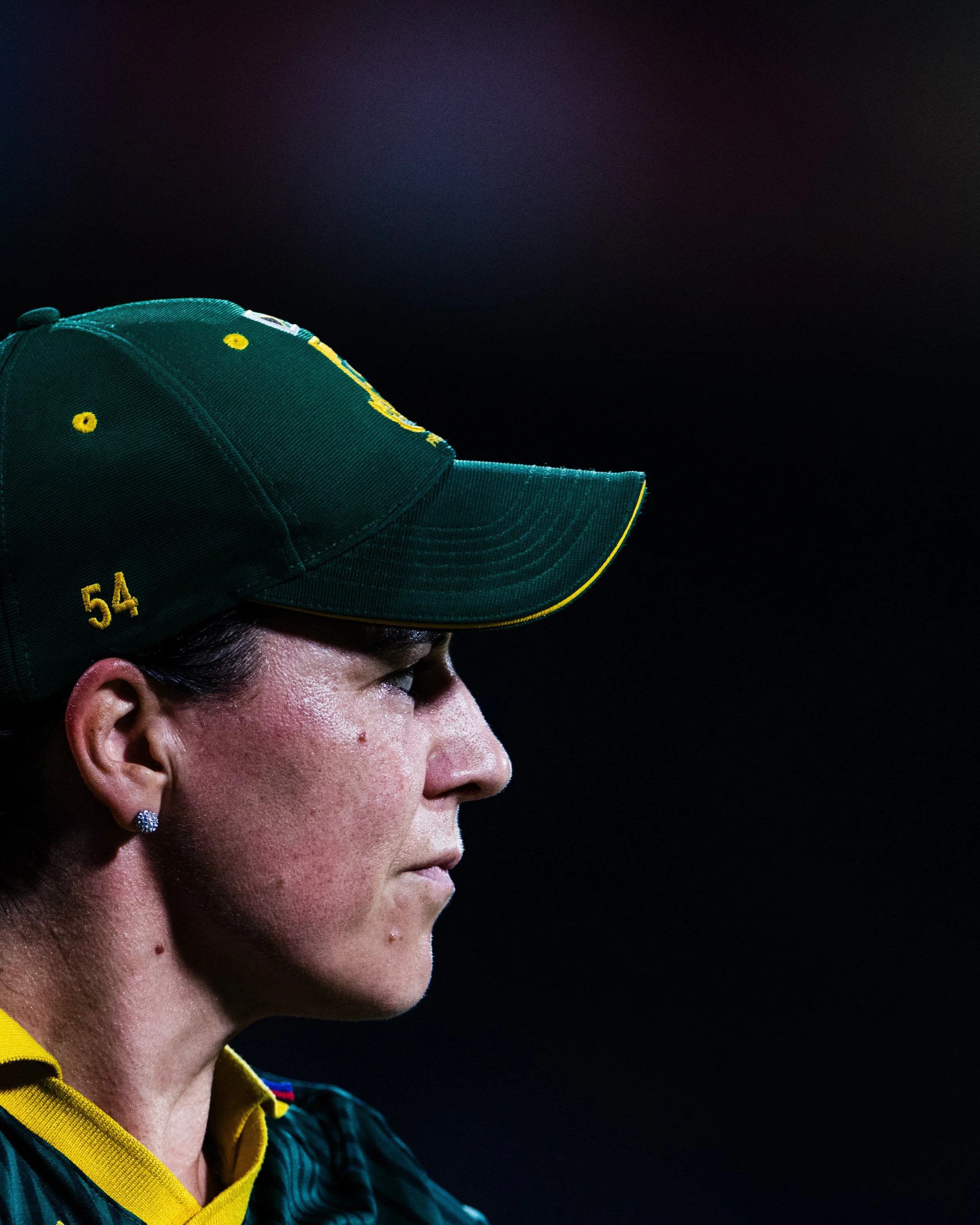 Close-up side profile of a woman in a dark-green cap with yellow details, wearing a yellow and black sports shirt, against a dark background.
