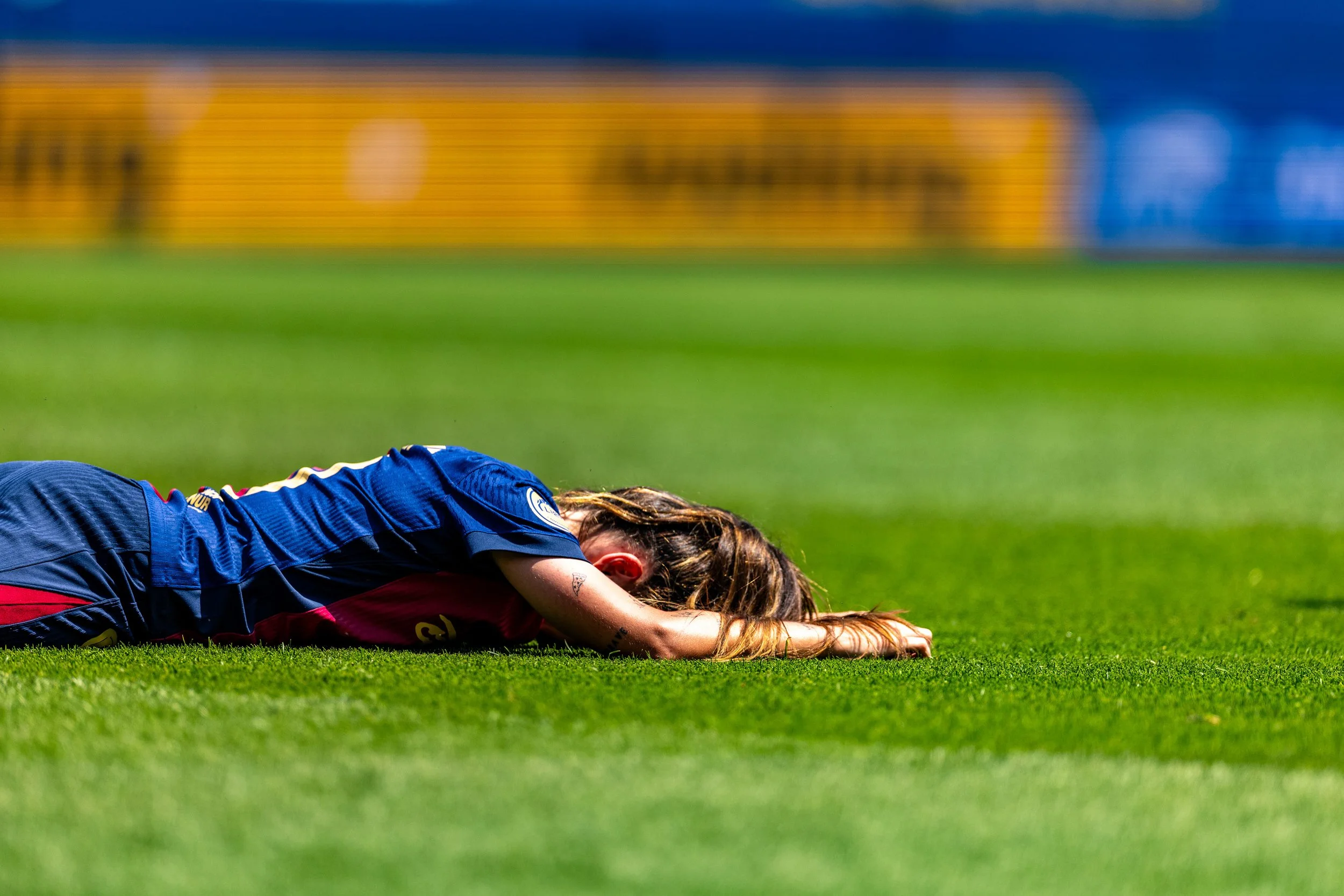A female soccer player is lying face down on the grass field, stretching or resting after a game, wearing a blue and red uniform.