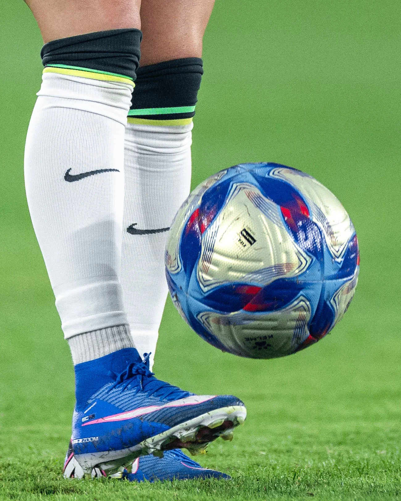 Close-up of a soccer player wearing blue sneakers, white socks, and black shorts with green and yellow accents, kicking a blue, white, and red soccer ball on a green field.