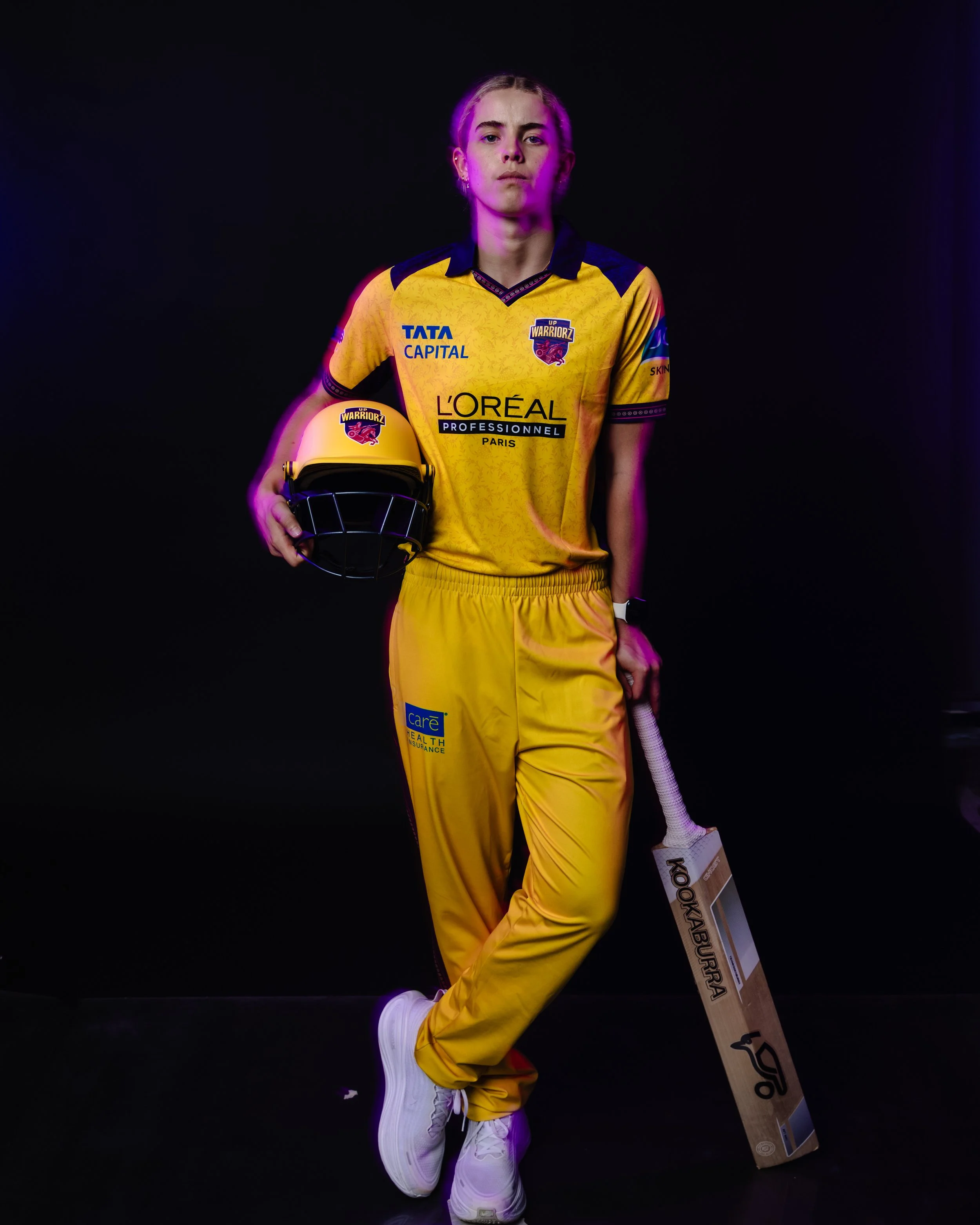 A young woman in a yellow sports jersey and yellow pants standing against a black background, holding a yellow helmet with a Warriors logo and a wooden cricket bat labeled 'Kookaburra.' She is wearing white sneakers and has blonde hair styled in brai