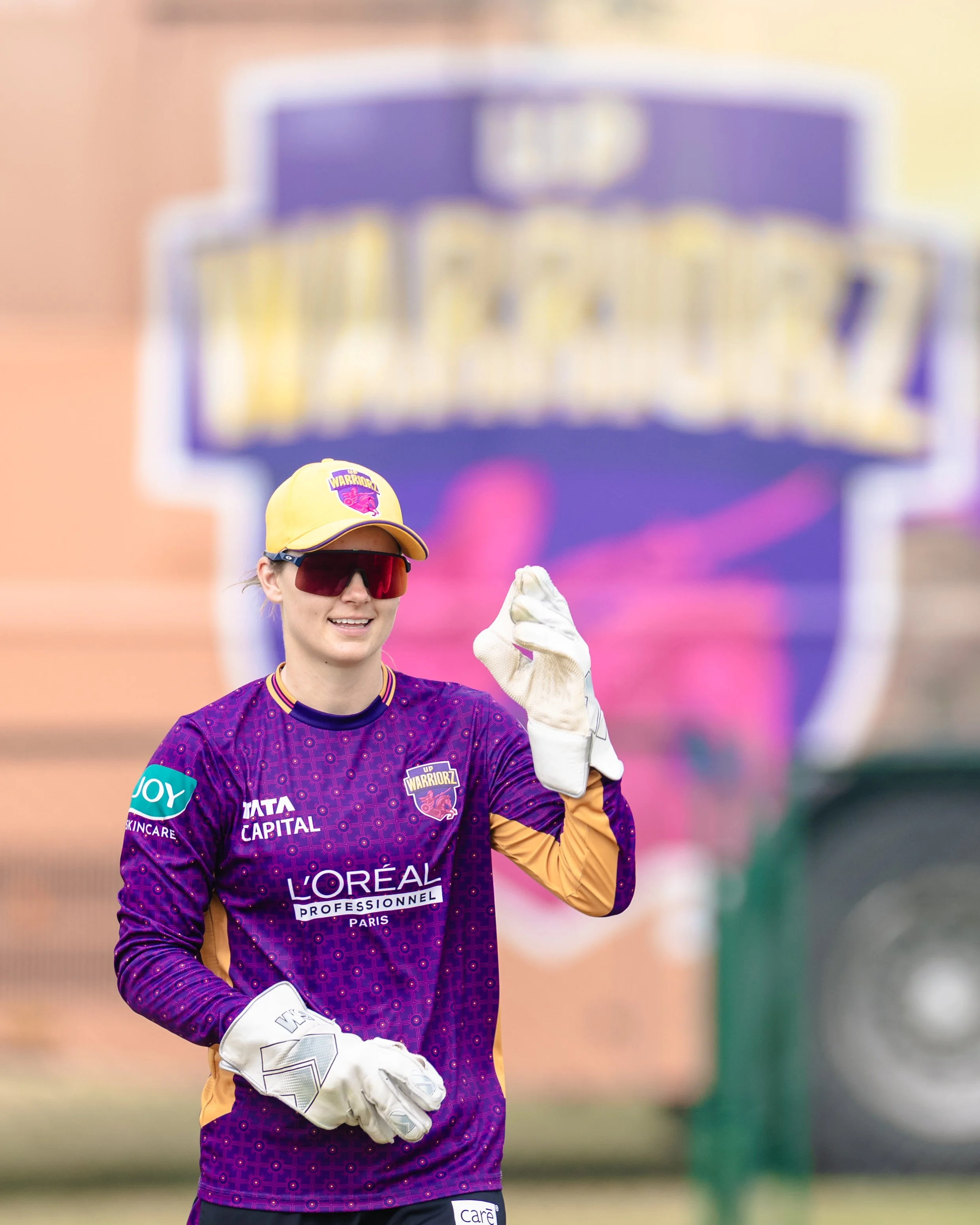 A female soccer player wearing a purple team jersey with sponsor logos, a yellow hat with a team logo, and sunglasses, holding a soccer ball and smiling, outdoors on a sports field.