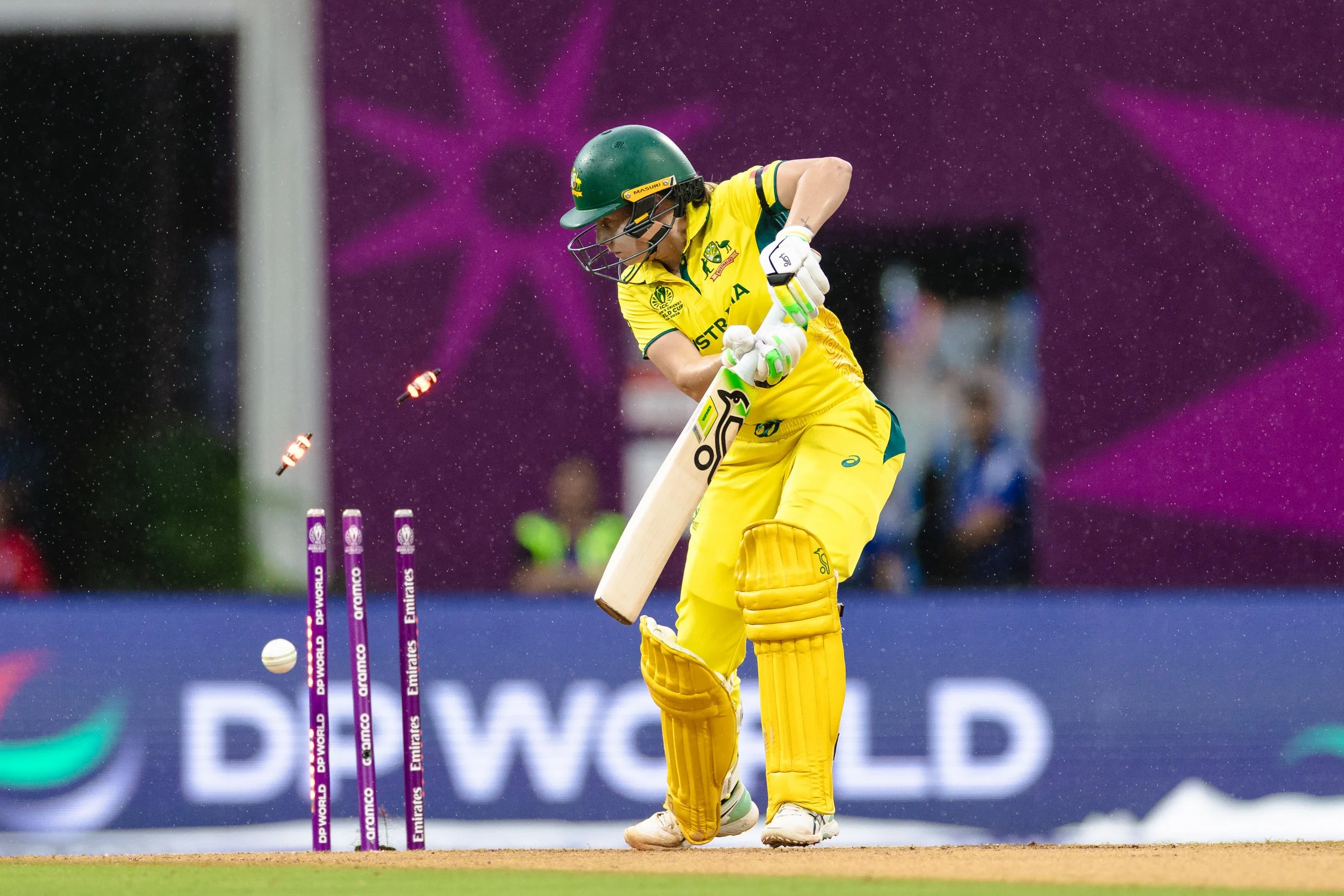 A cricket player in a yellow uniform and green helmet appears to be batting, with a cricket ball approaching and stumps in the background during a match.