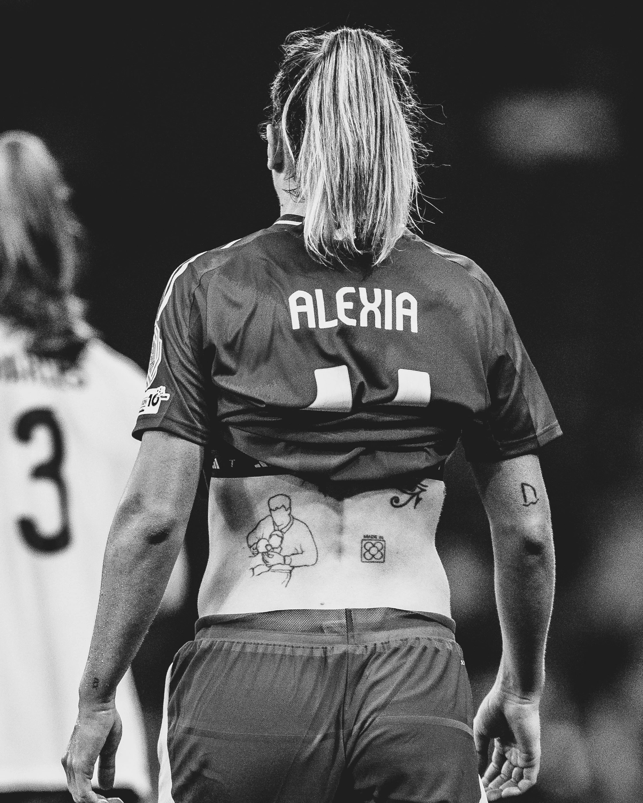 Back view of a female athlete with long blonde hair tied in a ponytail, wearing a sports jersey with the name 'ALEXIA' on the back and sports shorts, standing on a sports field.