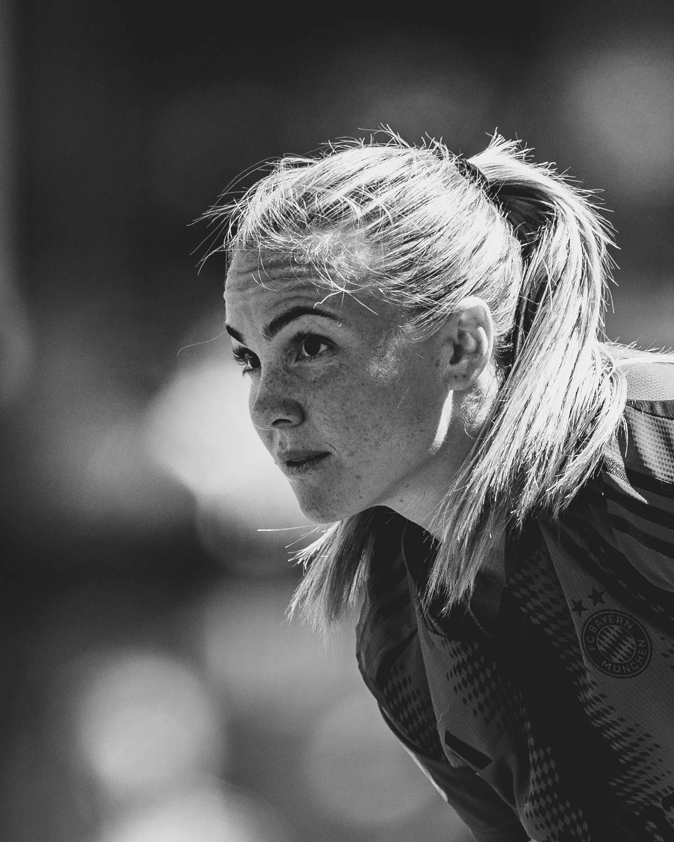 Close-up of a female athlete with blonde hair tied back, wearing a sports jersey with a Bayern Munich logo, in black and white with a focused expression.