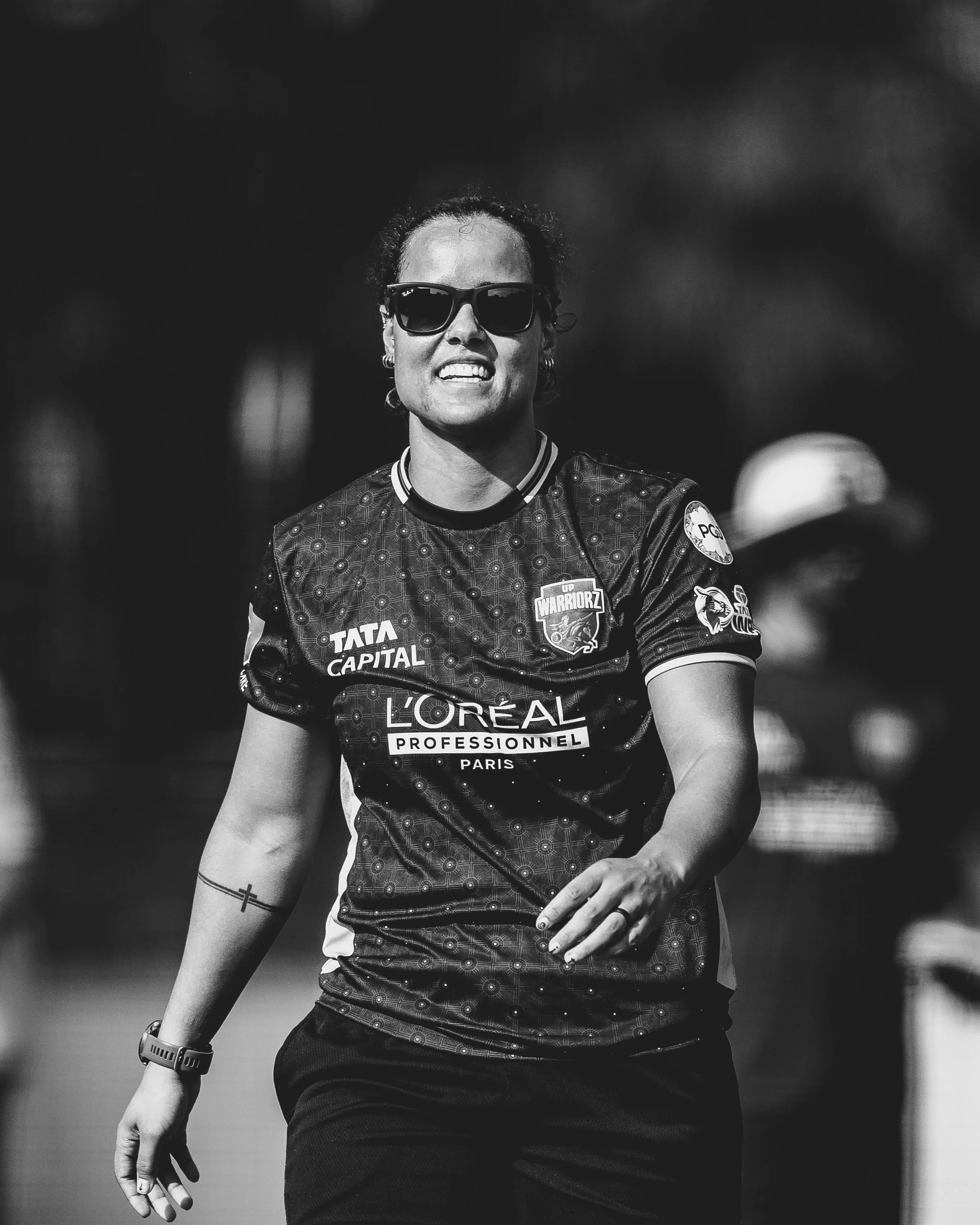 A woman wearing sunglasses and a sports uniform with sponsorship logos, including Tata Capital and L'Oreal, is shown in a black and white photograph.