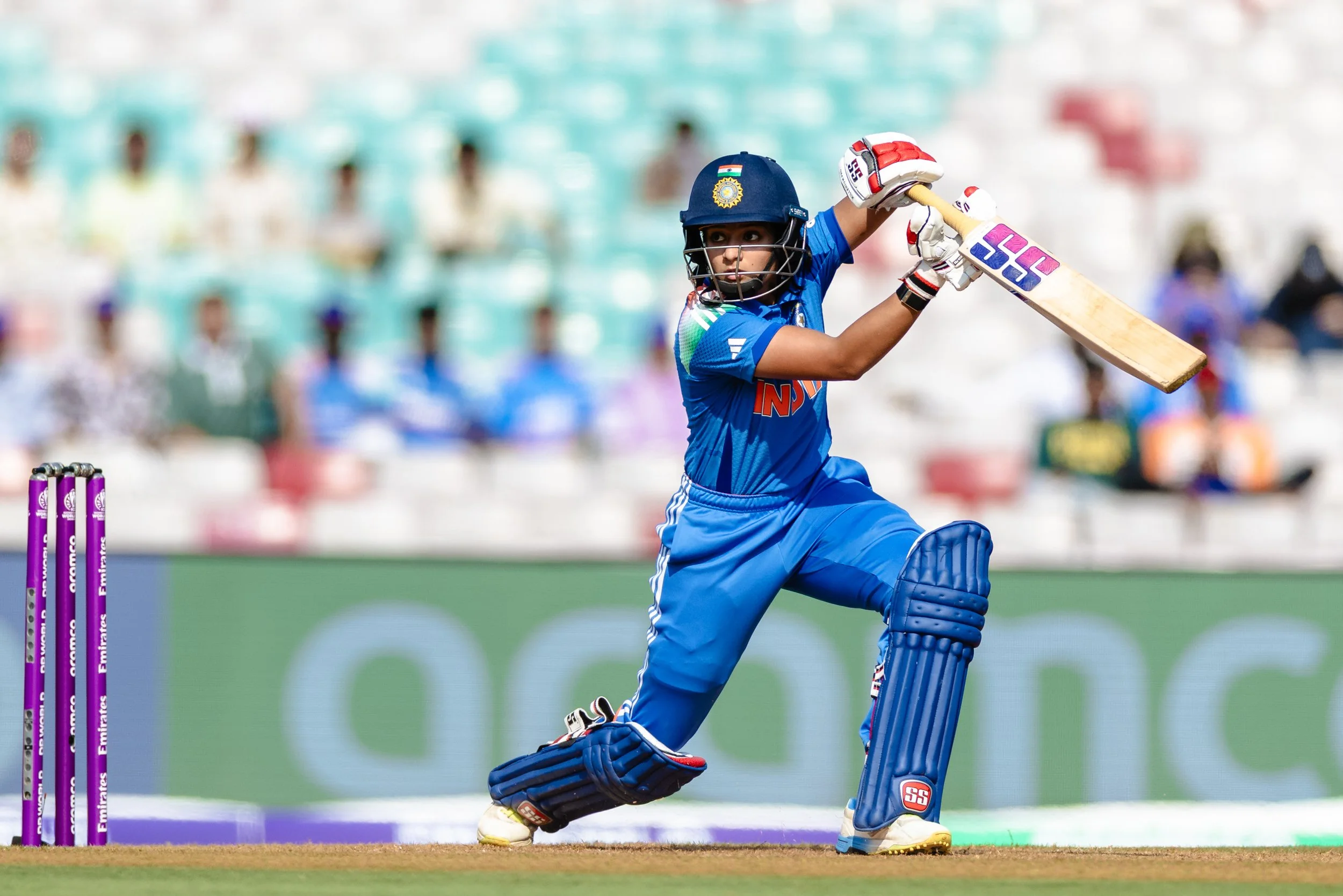 A young girl playing cricket, wearing a blue sports uniform and protective gear, holding a cricket bat, in a stadium with blurred spectators in the background.