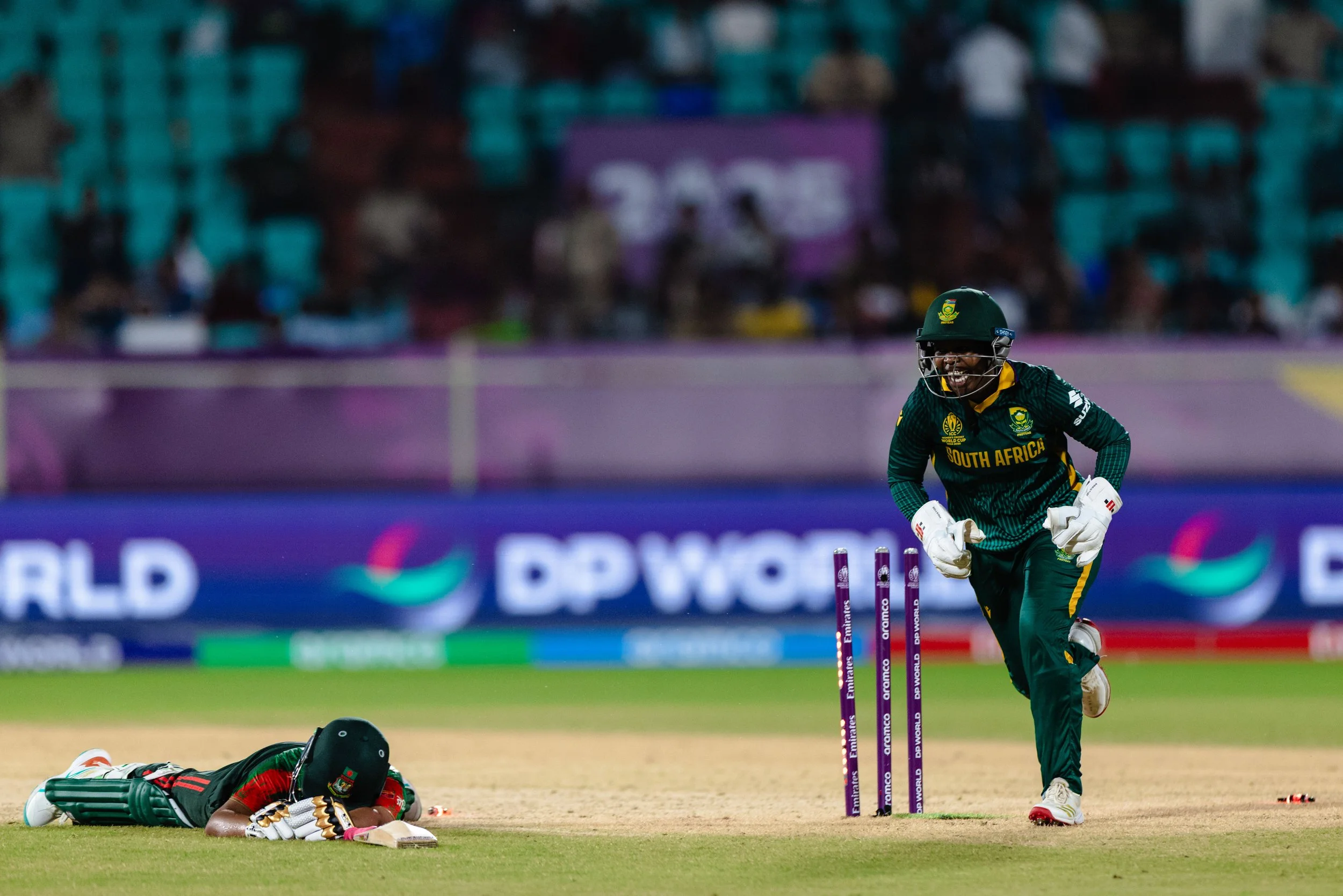A cricket player from South Africa celebrating after hitting the stumps, while a player from Bangladesh lies on the ground. The game takes place on a cricket field with a crowd in the background.