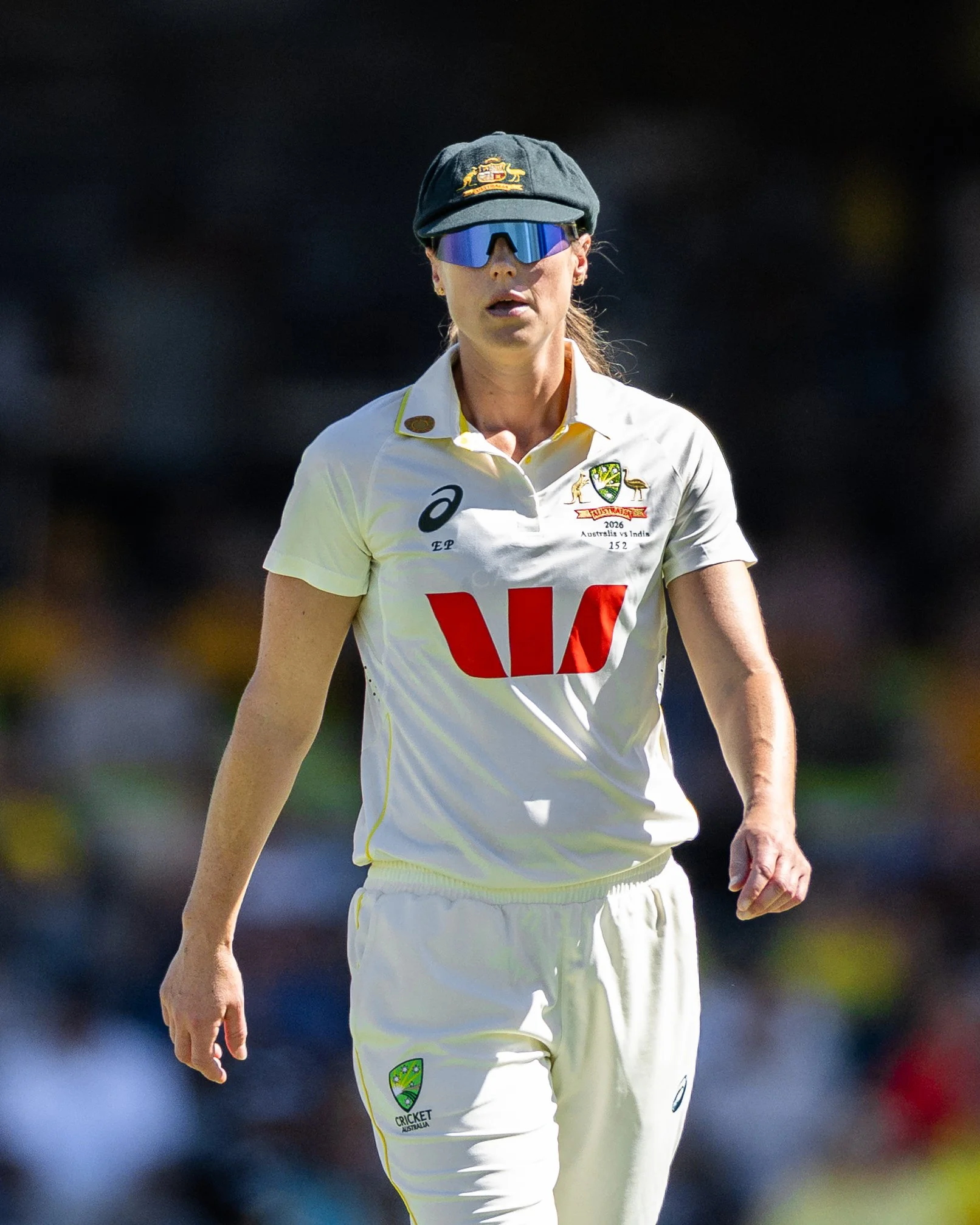 Female cricket player in white uniform with Australian emblem, wearing a black cap and sunglasses, on the cricket field.