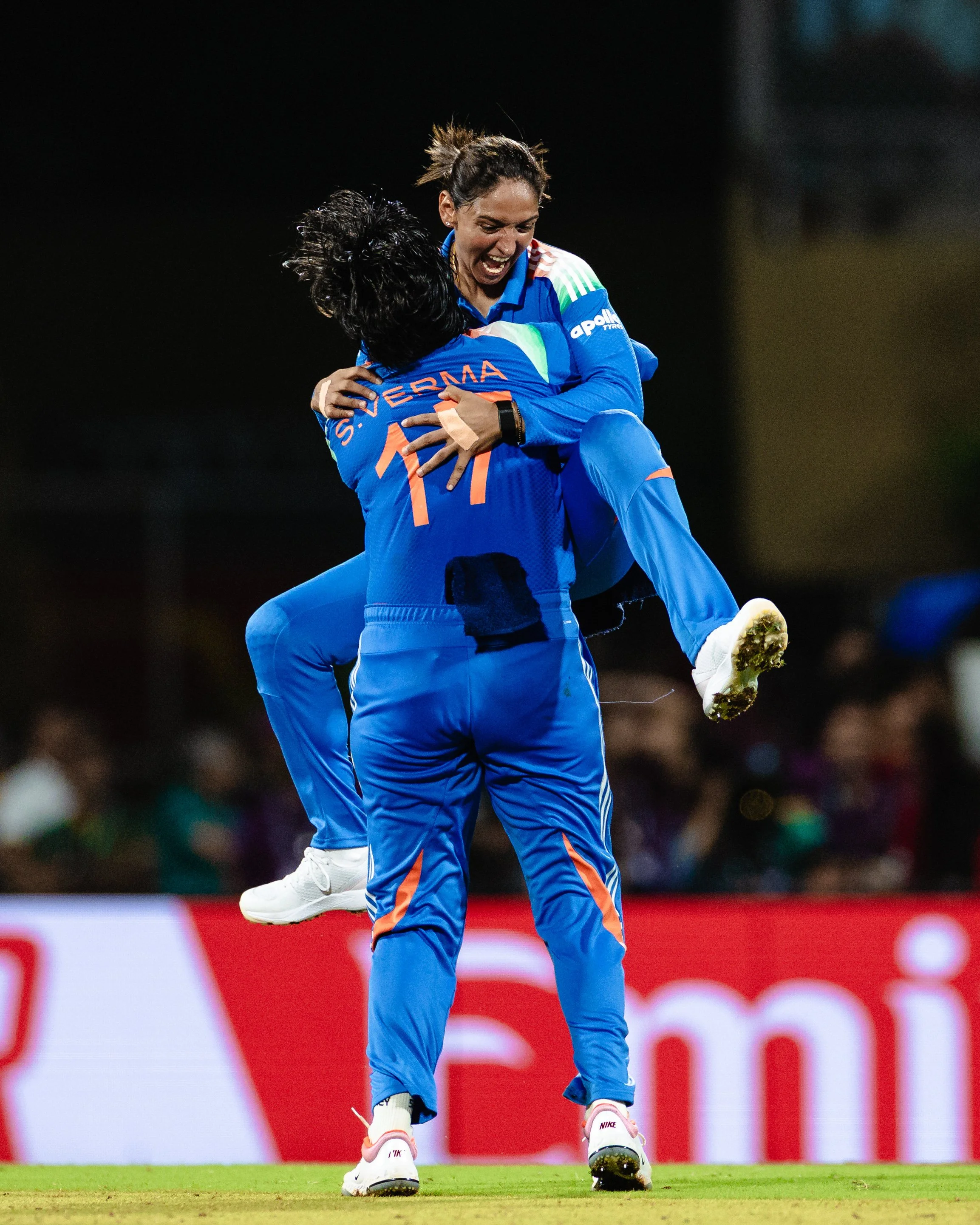 Two female cricket players in blue uniforms celebrating on the field. One is lifting the other in a joyful embrace. The player being lifted has her head down and arms around her teammate, while the other is smiling broadly. They are standing on green