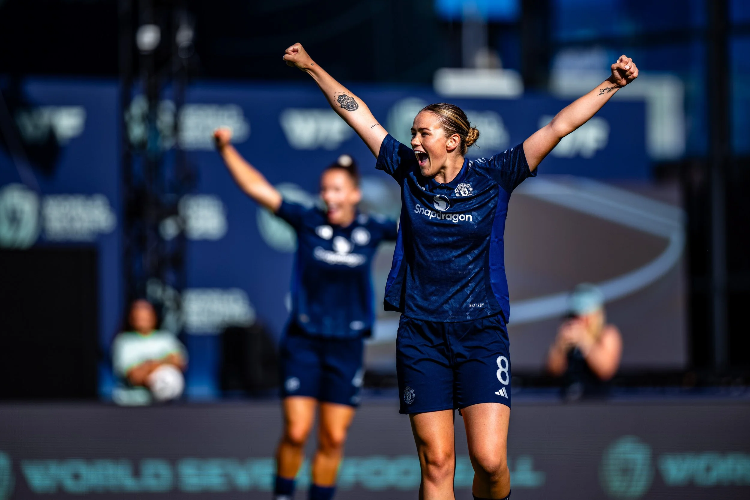 A female soccer player celebrating with arms raised on the field, wearing a navy blue jersey and shorts, with tattoos on her arms and a smile on her face. Other players are in the background.