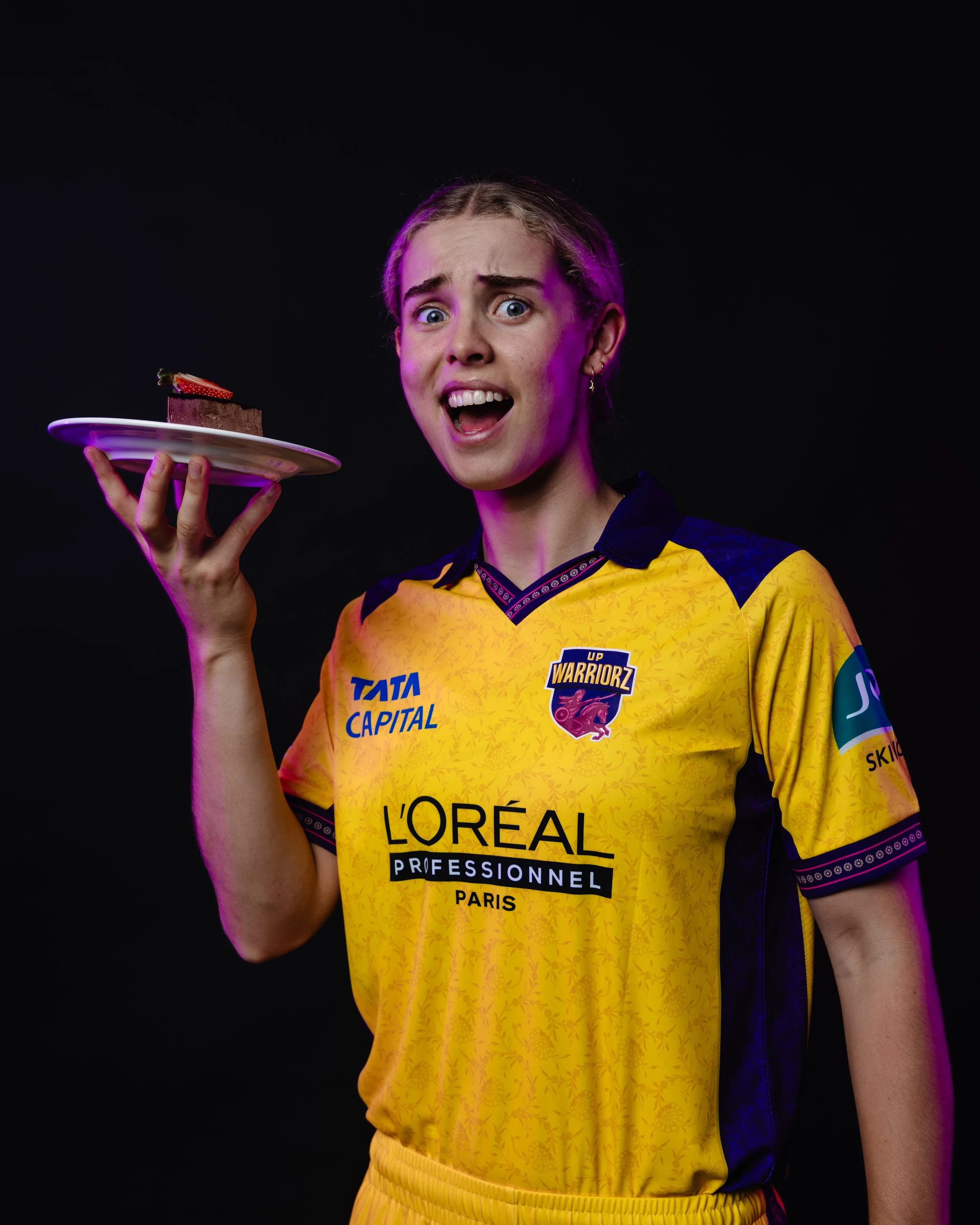A woman wearing a yellow sports jersey with various sponsors, holding a white plate with a slice of chocolate cake topped with a strawberry, against a black background.