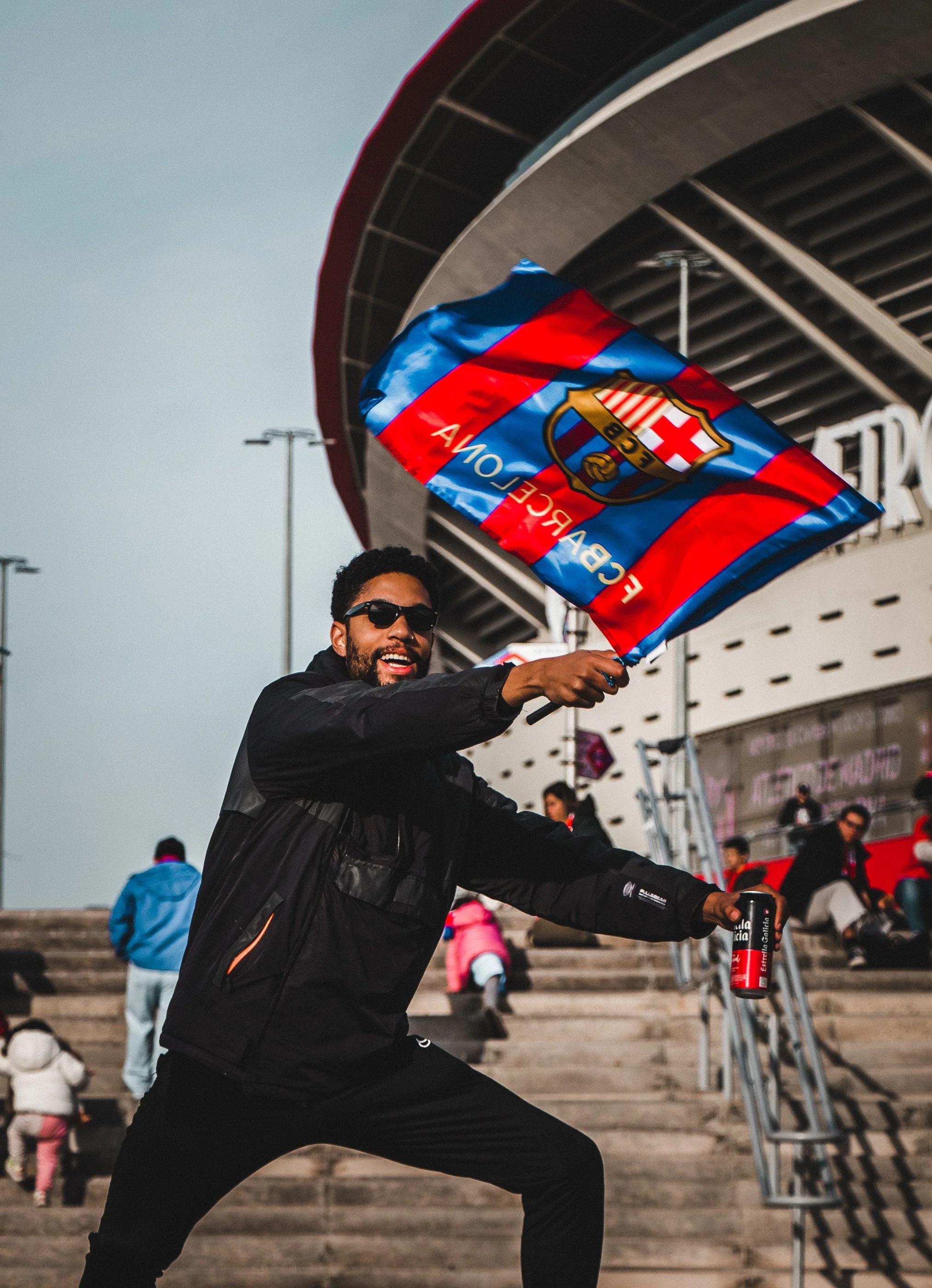 A man with sunglasses smiling, holding a flag with the FC Barcelona logo, standing on steps outside a stadium with other people in the background.