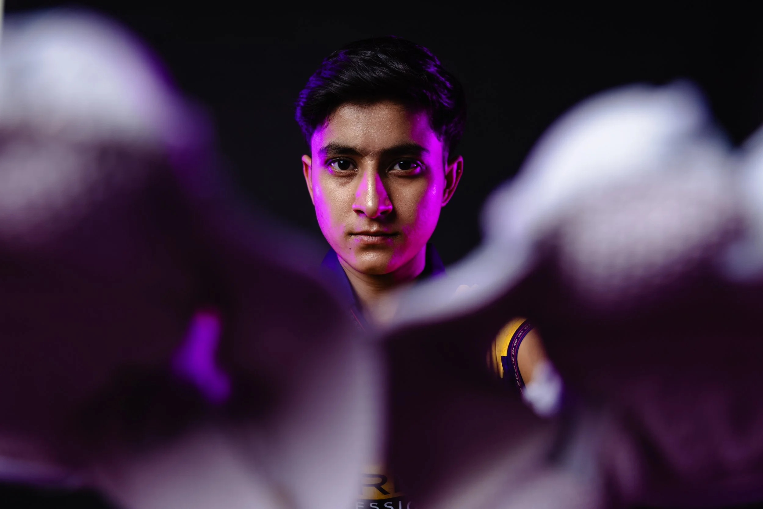 Young man with short black hair looking directly at the camera, with a dark background and purple lighting highlighting his face.
