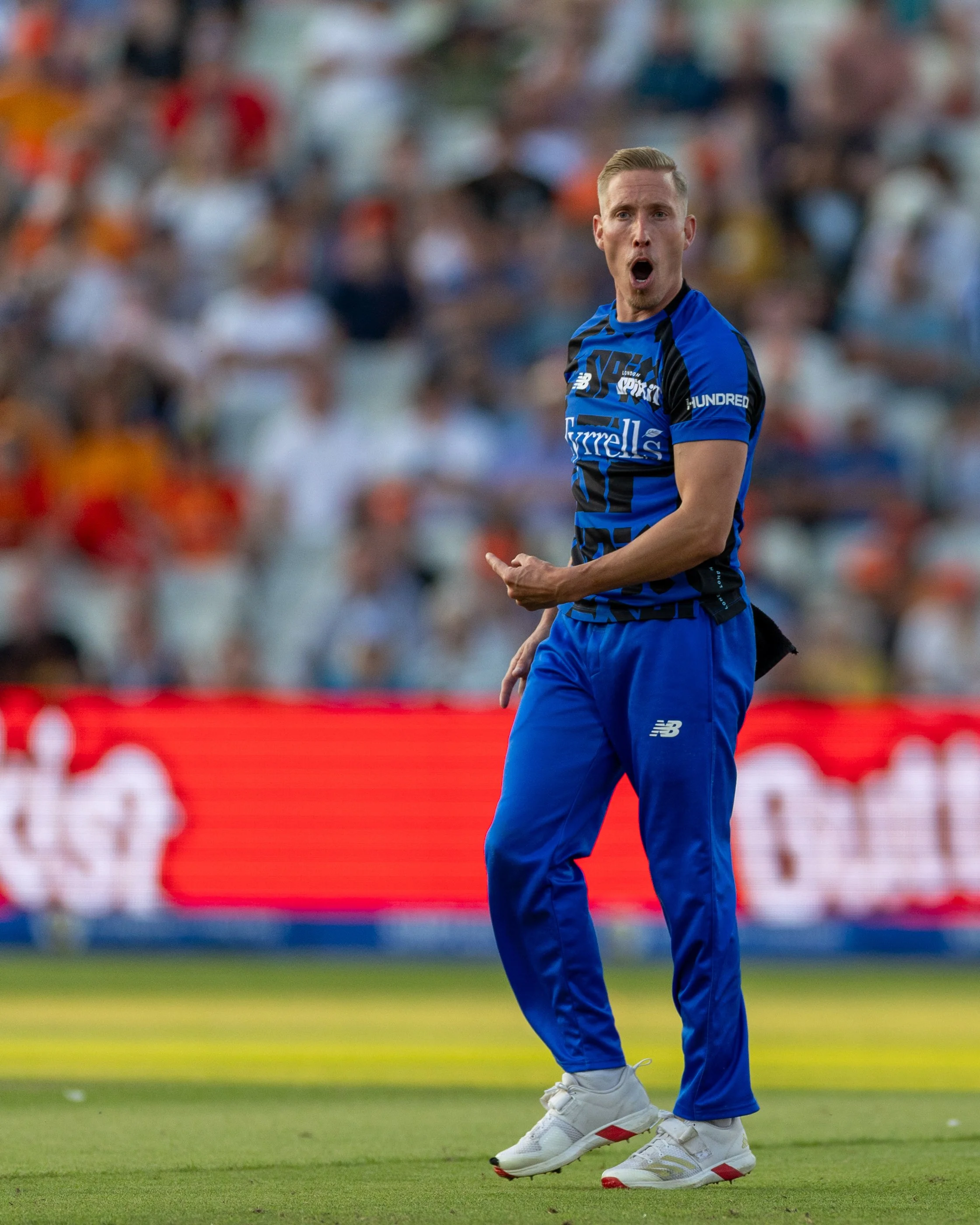 A man in a blue sports jersey and pants, wearing white sneakers, is on a cricket field during a match, with a blurred crowd in the background, celebrating or reacting to a play.