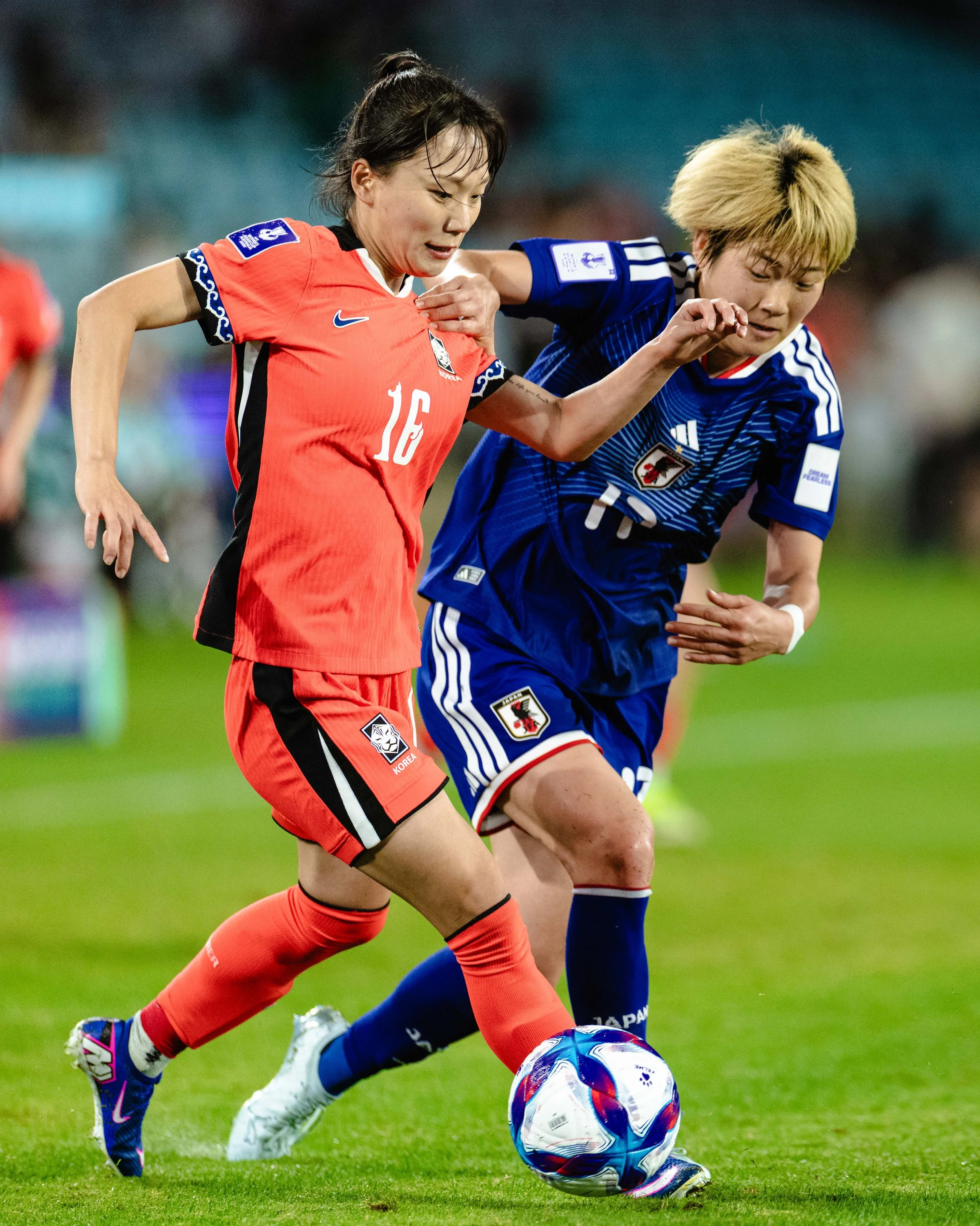 Two women playing soccer on a field, one in a red jersey with the number 16 and the other in a blue jersey, competing for the ball.