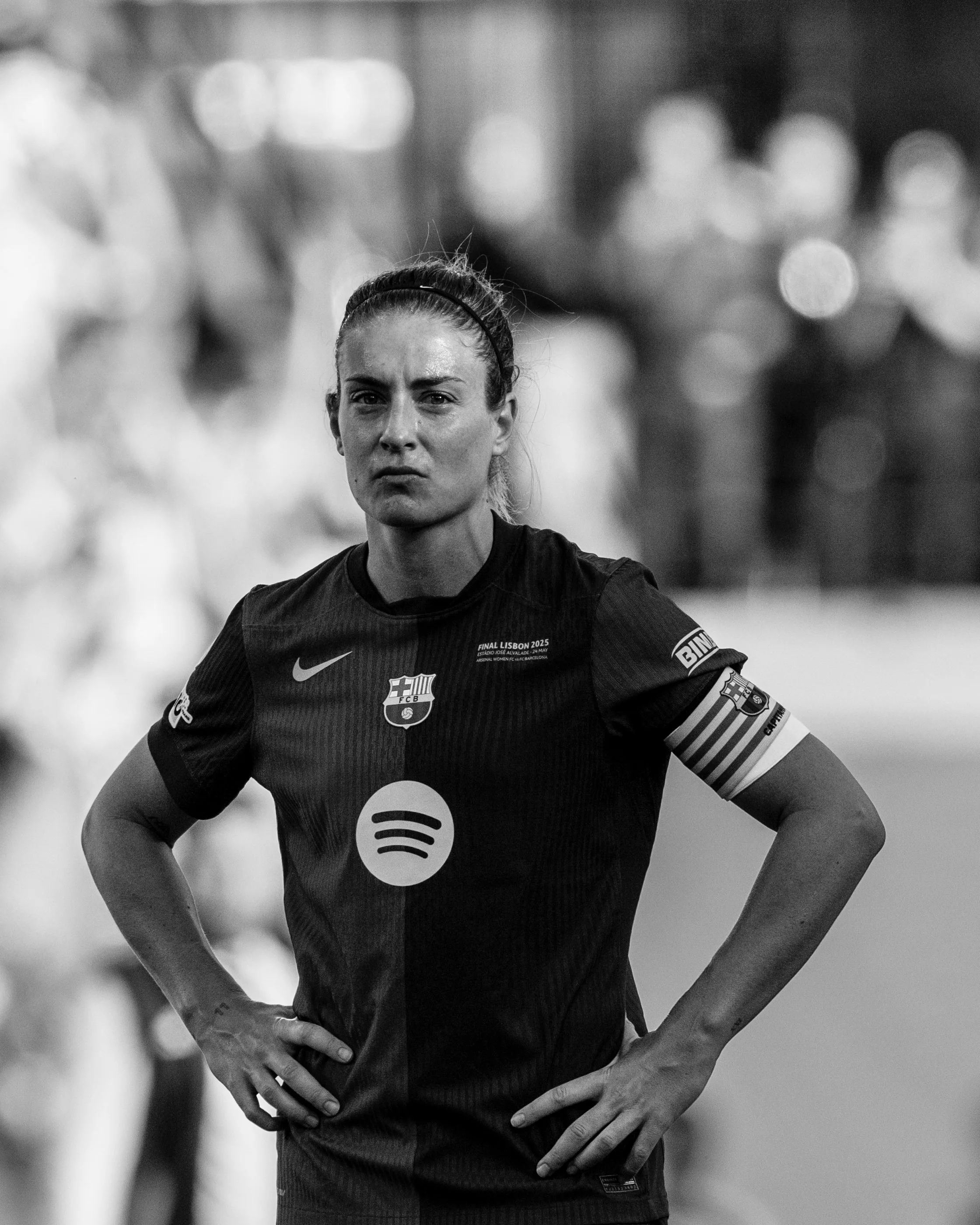 A female soccer player with her hands on her hips, wearing a black jersey with sponsor logos and a captain's armband, standing on a field with a blurred crowd in the background.