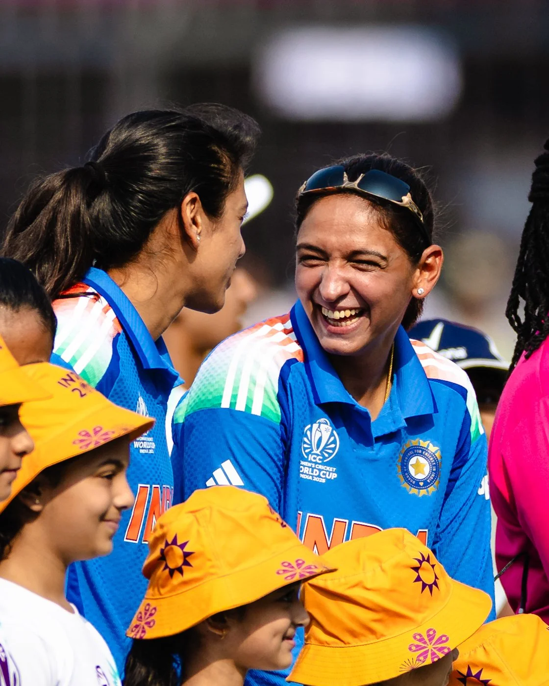 Indian women cricket players wearing blue jerseys and Indian children in yellow hats, smiling and talking during a cricket event.