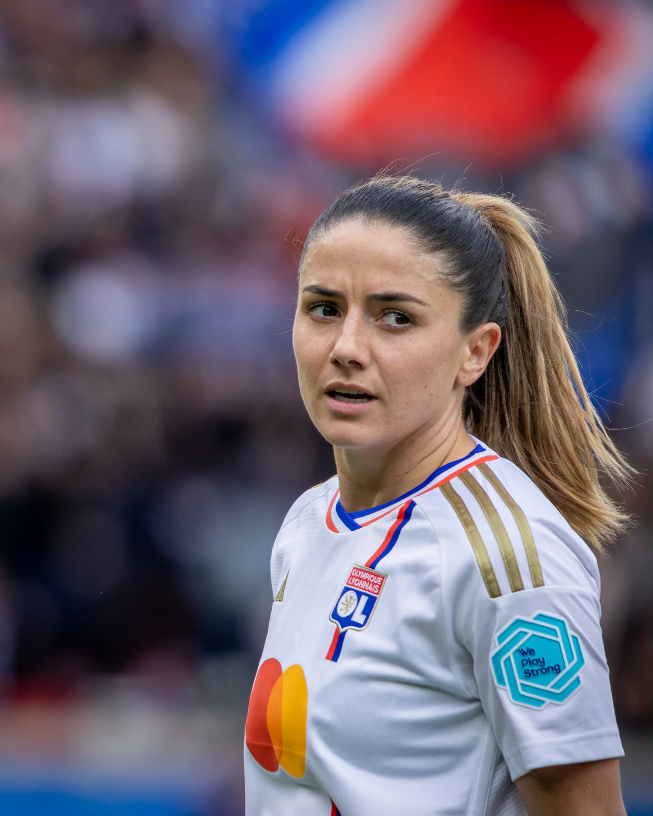 A female soccer player with a ponytail, wearing a white jersey with blue, red, and gold accents, and logos including "Olympique Lyonnais" and "We Play Strong."