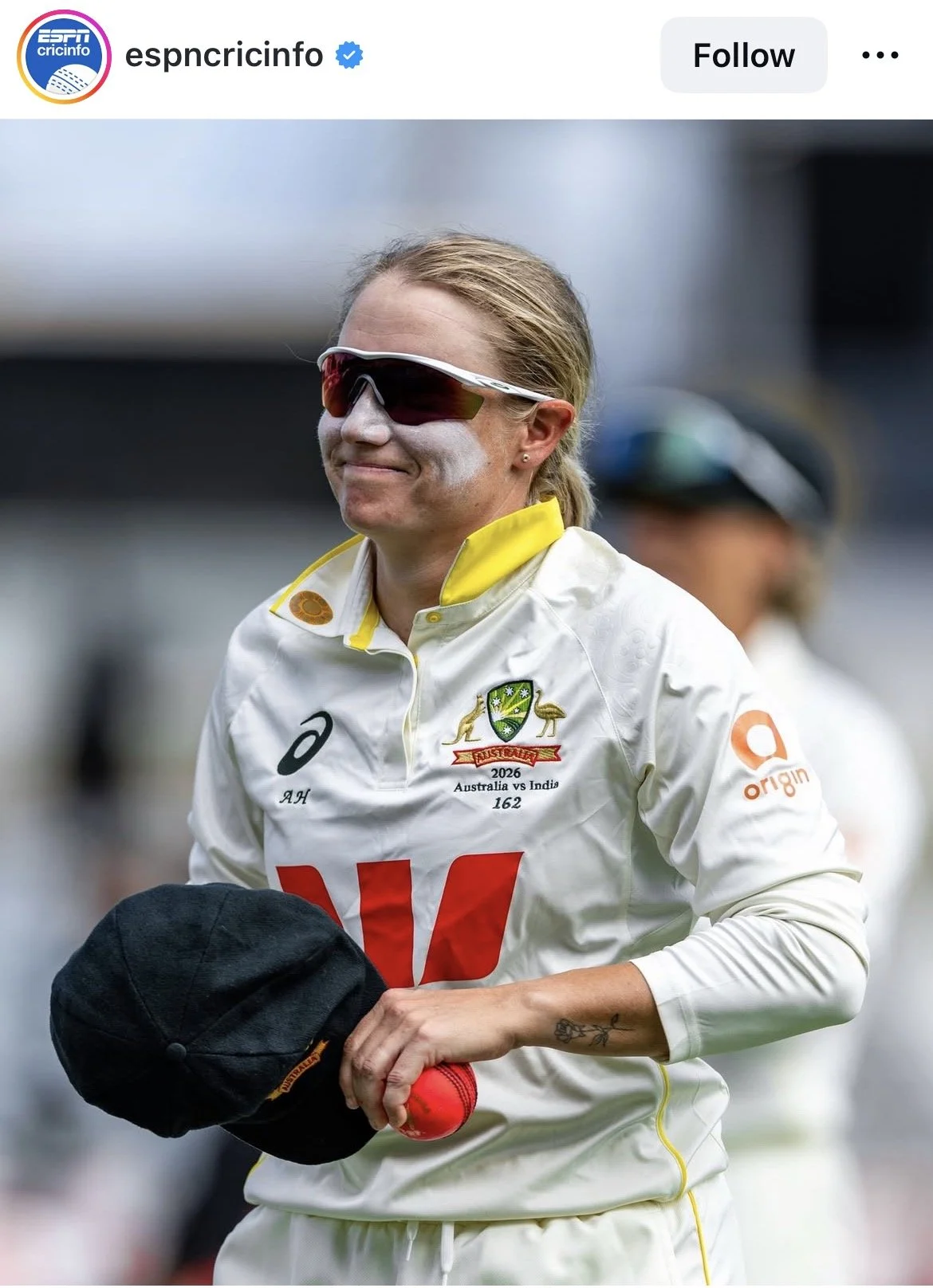 Female cricketer wearing white Australia team uniform with face paint, holding a black cap and red cricket ball, wearing sports sunglasses, standing outdoors.