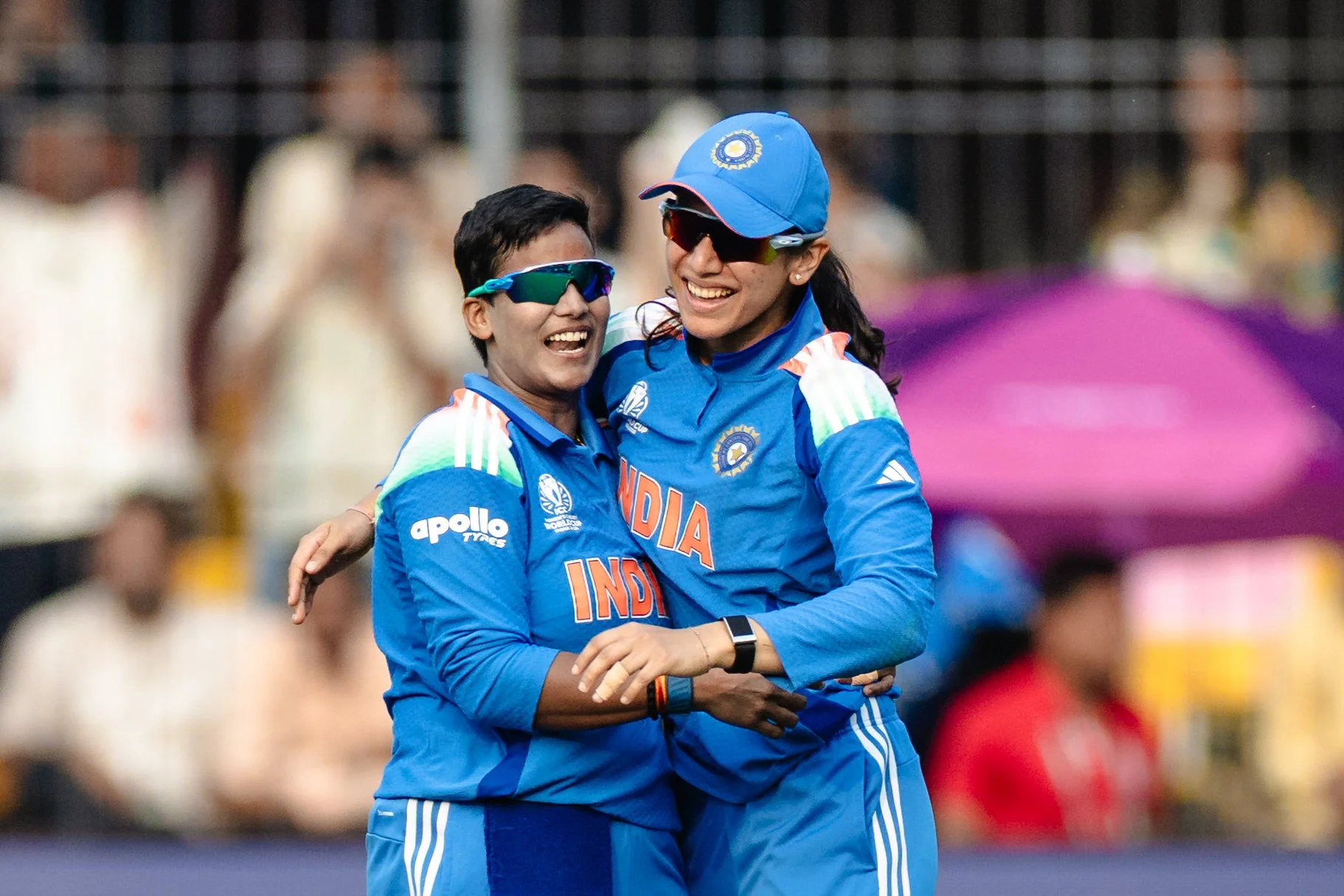 Two Indian female cricket players in blue uniforms hugging and smiling at a cricket stadium.