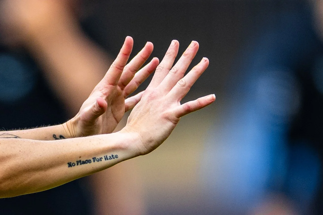 Close-up of two hands reaching out, one with a tattoo that says 'No Place For Hate' on the forearm.
