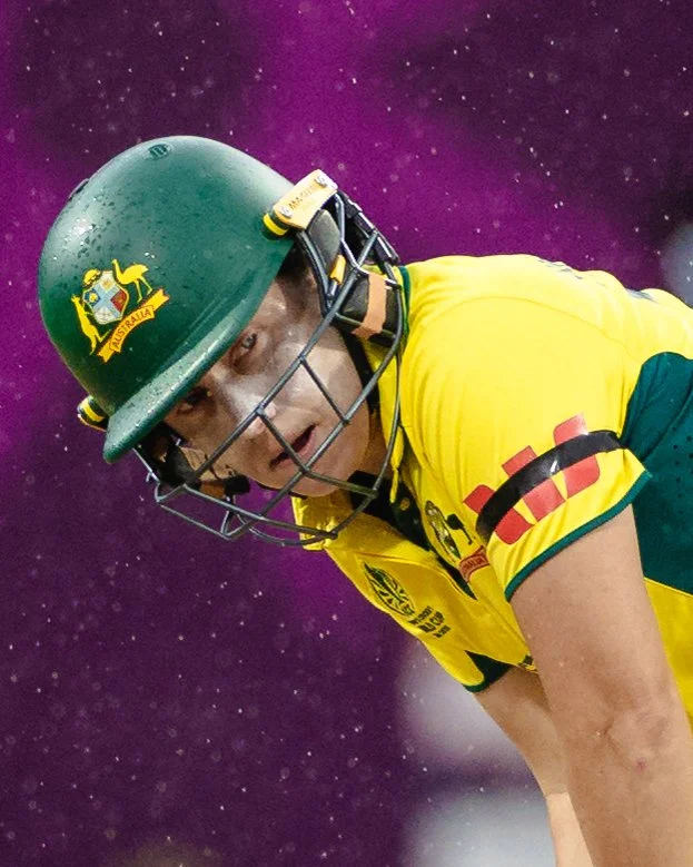 A cricket player in a yellow and green uniform wearing a green helmet with the Australian emblem, crouching down on a purple background with star-like patterns.