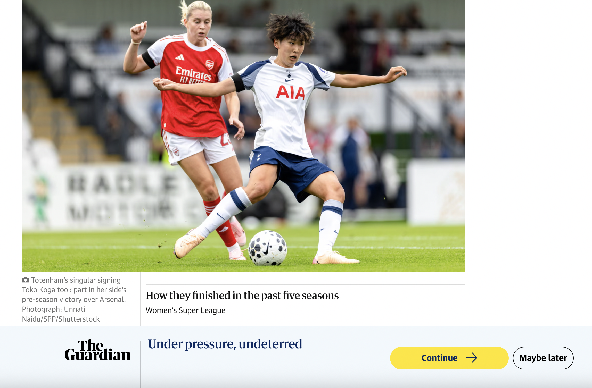 A soccer match showing two female players, one in a white and navy blue uniform and the other in a red and white uniform, competing for the ball on the field. The player in white is kicking the ball while the player in red is closely behind her.