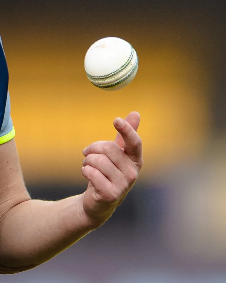 Close-up of a person’s hand with a bent elbow, balancing a white cricket ball with green and black stitching, against a blurred yellowish background.