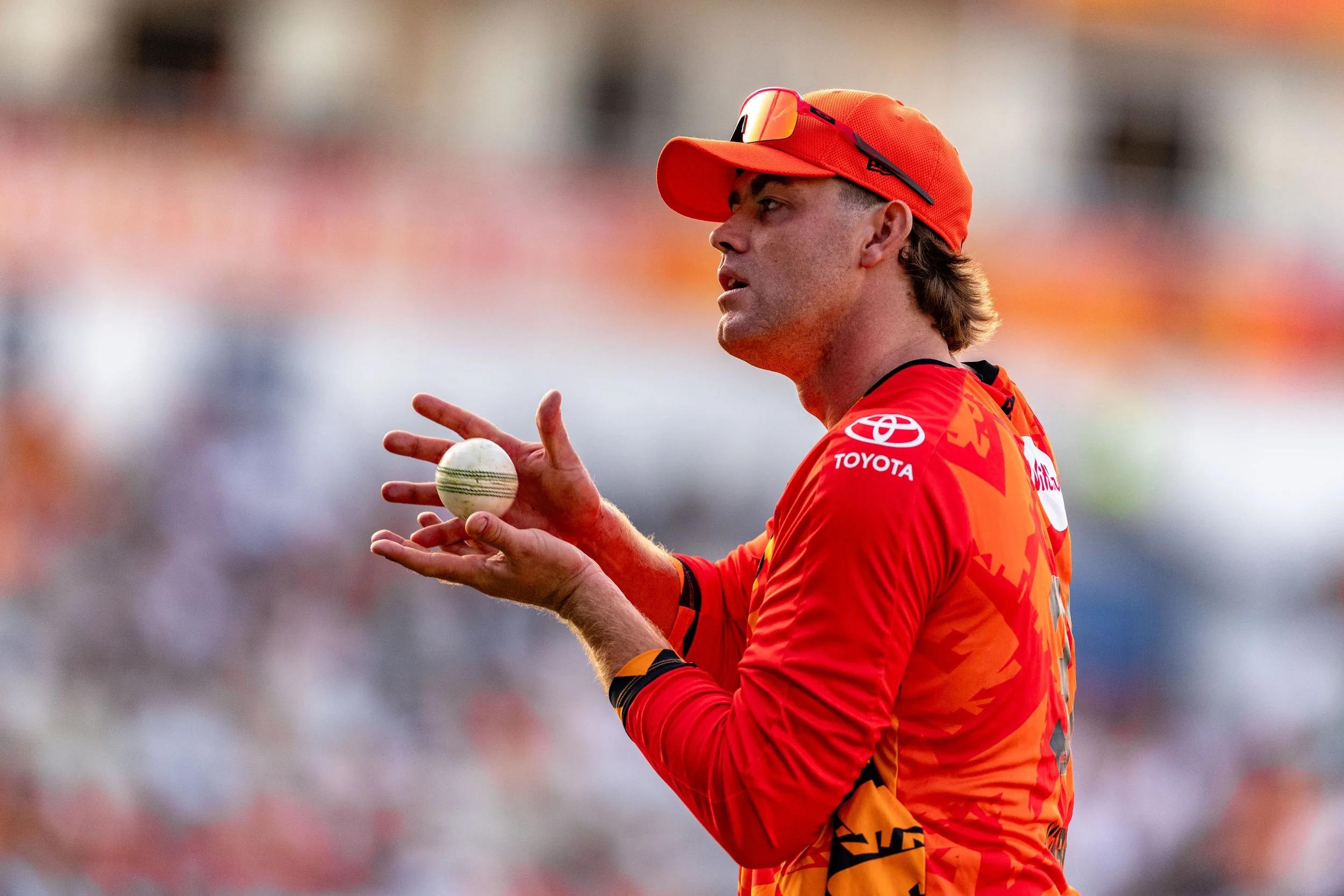 A cricket player wearing a red and orange uniform and cap, holding a cricket ball with one hand amid a blurred background.