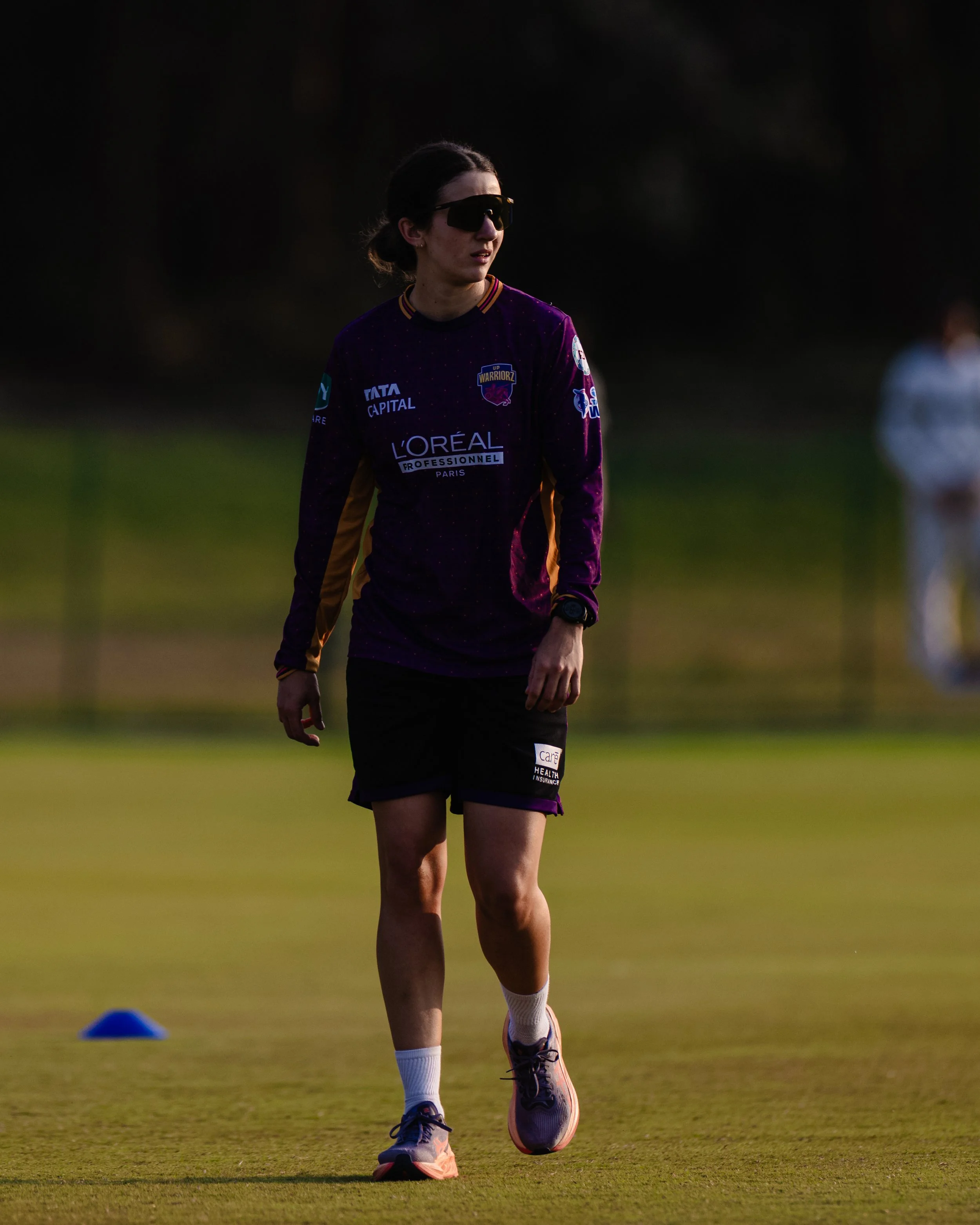 A woman in a purple sports jersey and black shorts walks on a green field during daytime, with a small blue cone on the ground nearby and another person in the background.