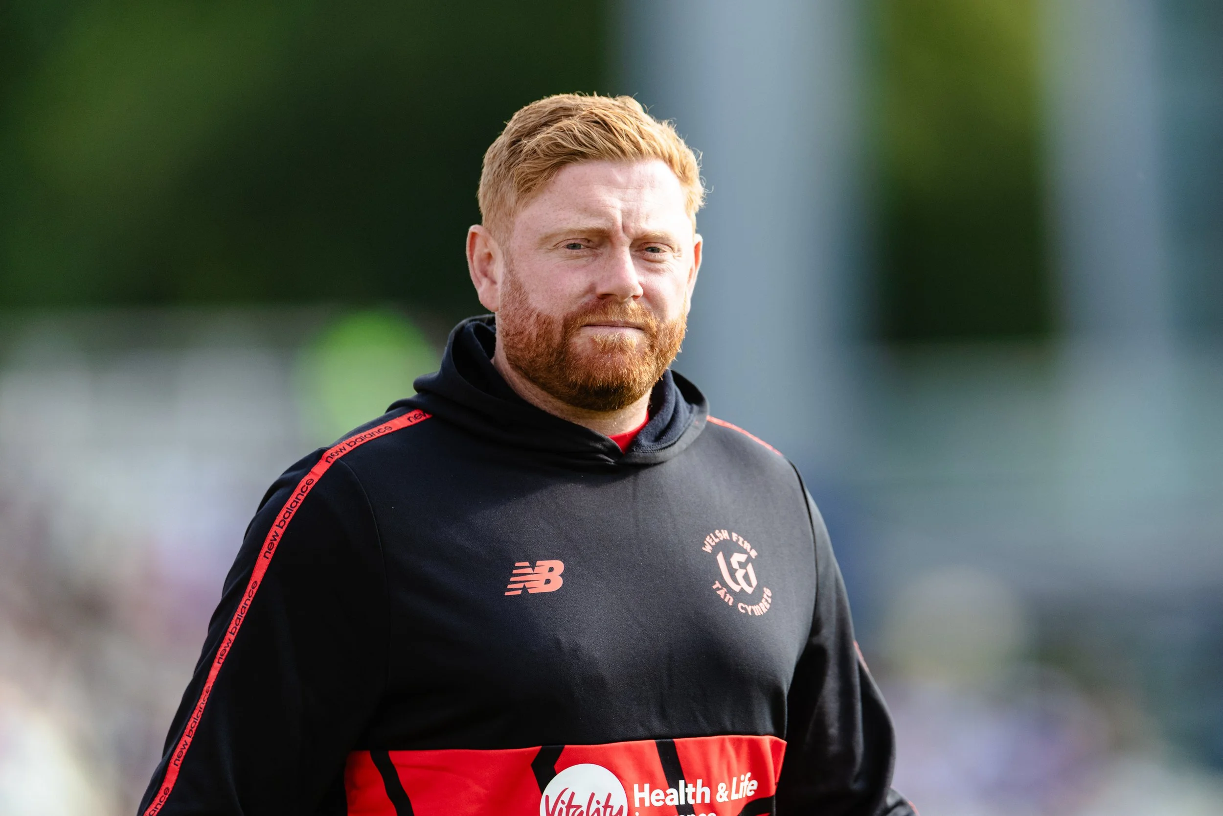 A man with red hair and beard wearing a black sports jacket with red accents, standing outdoors with a blurred green background.