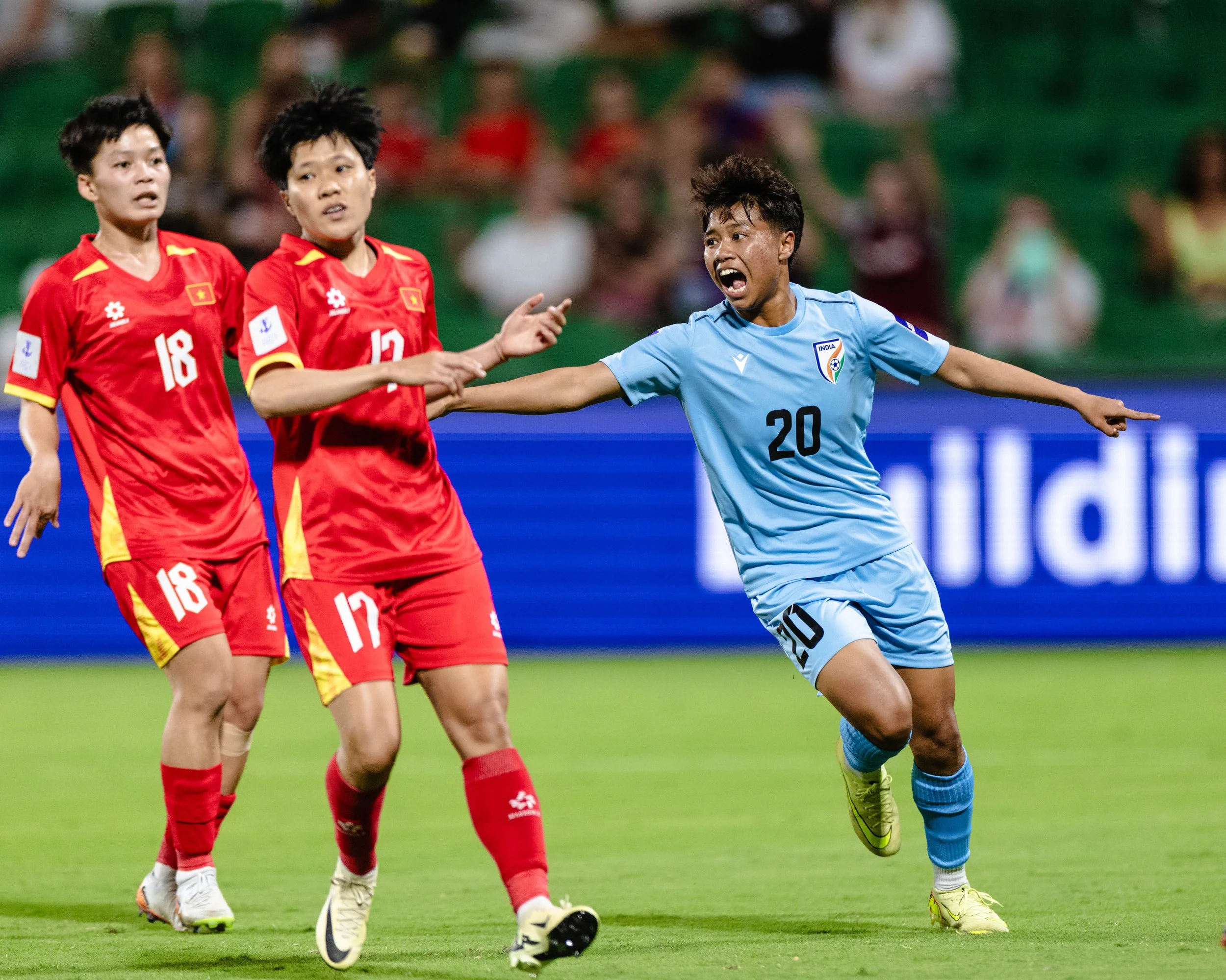 A soccer match with four female players, three in red uniforms and one in a light blue uniform, competing on a green field with a blurred audience in the background.